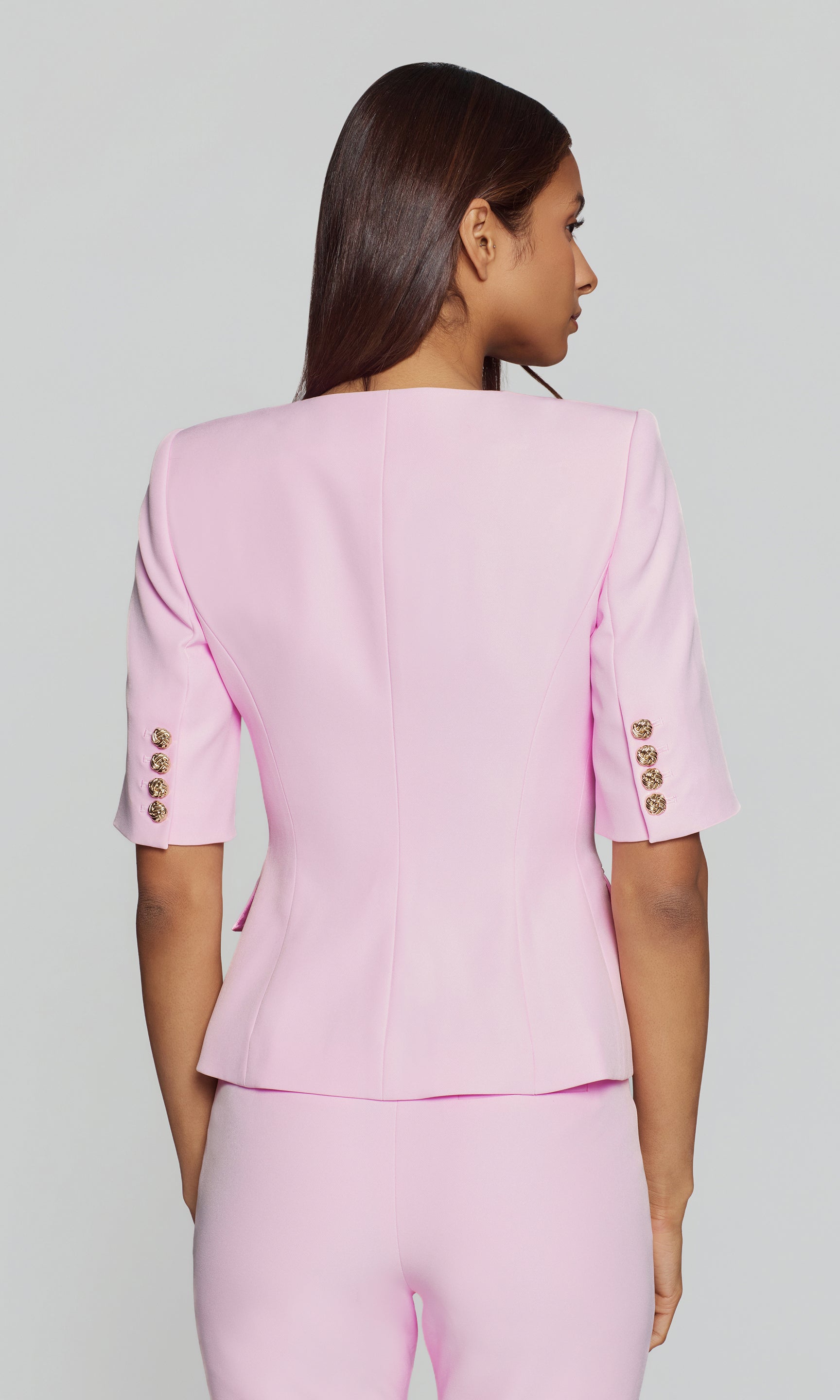 Back view of woman wearing a pink outfit with gold buttons on a gray background