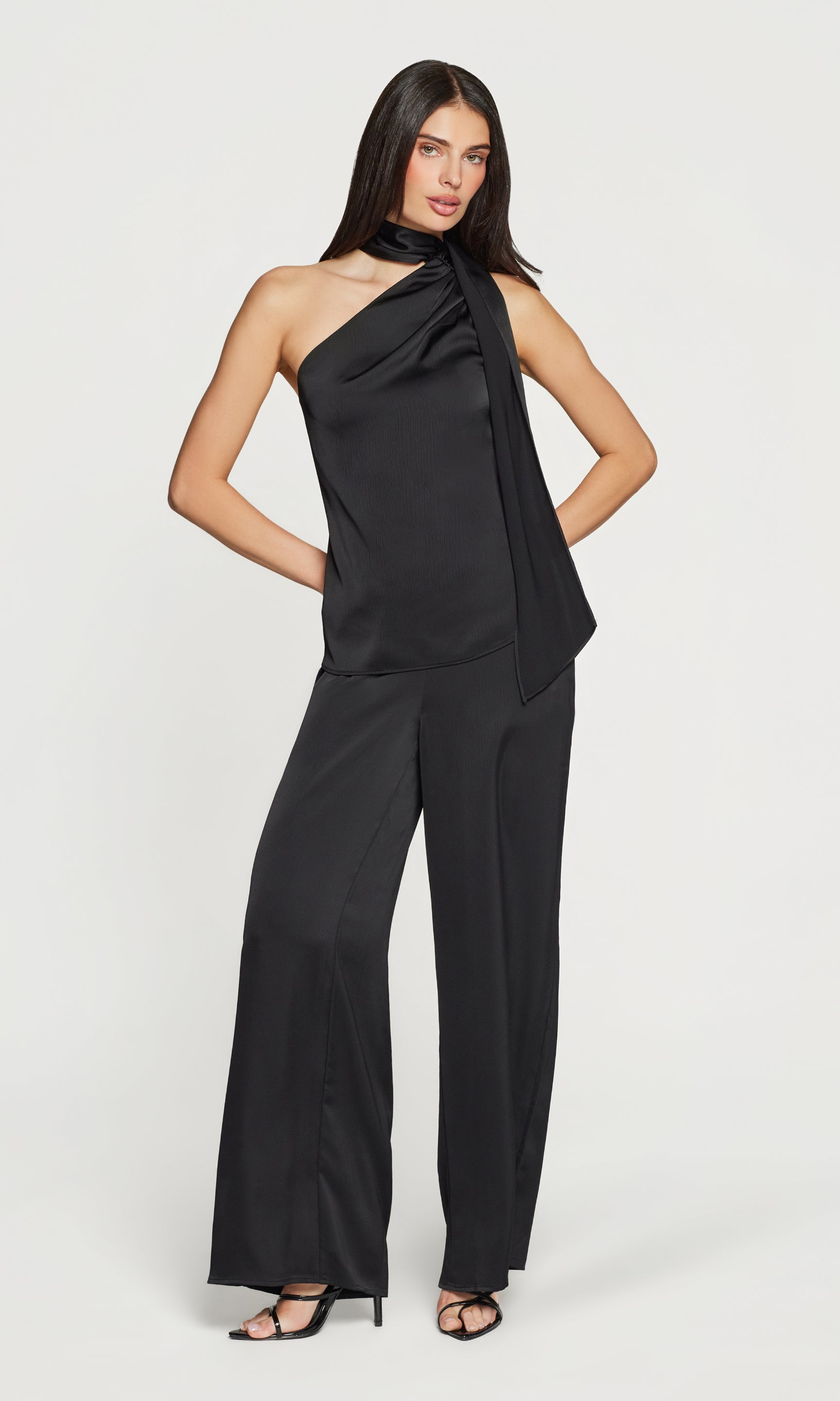 Woman wearing a black one-shoulder top and matching pants with heels on a white background