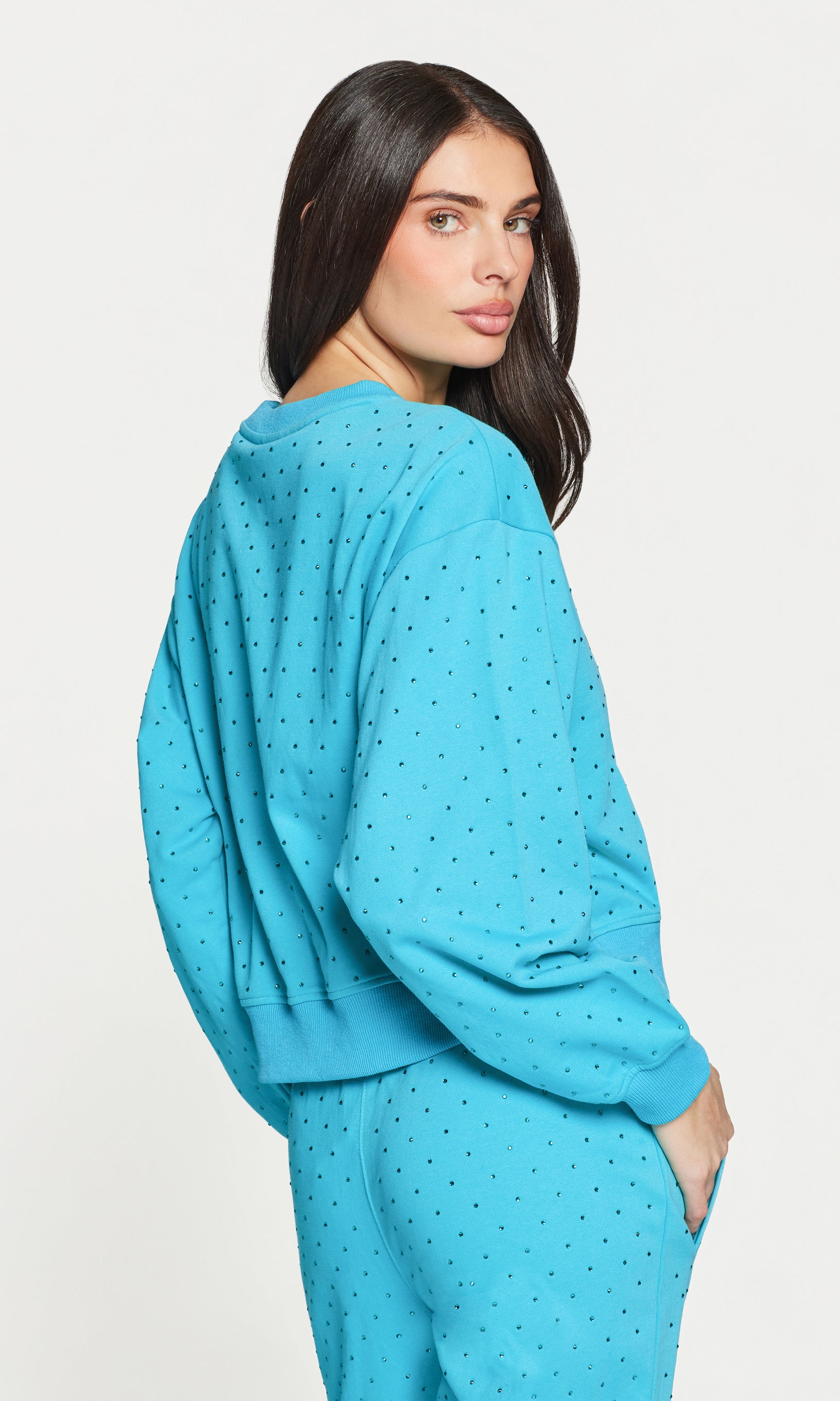 3/4 back view of woman wearing a blue crystal embellished sweater on a light gray background
