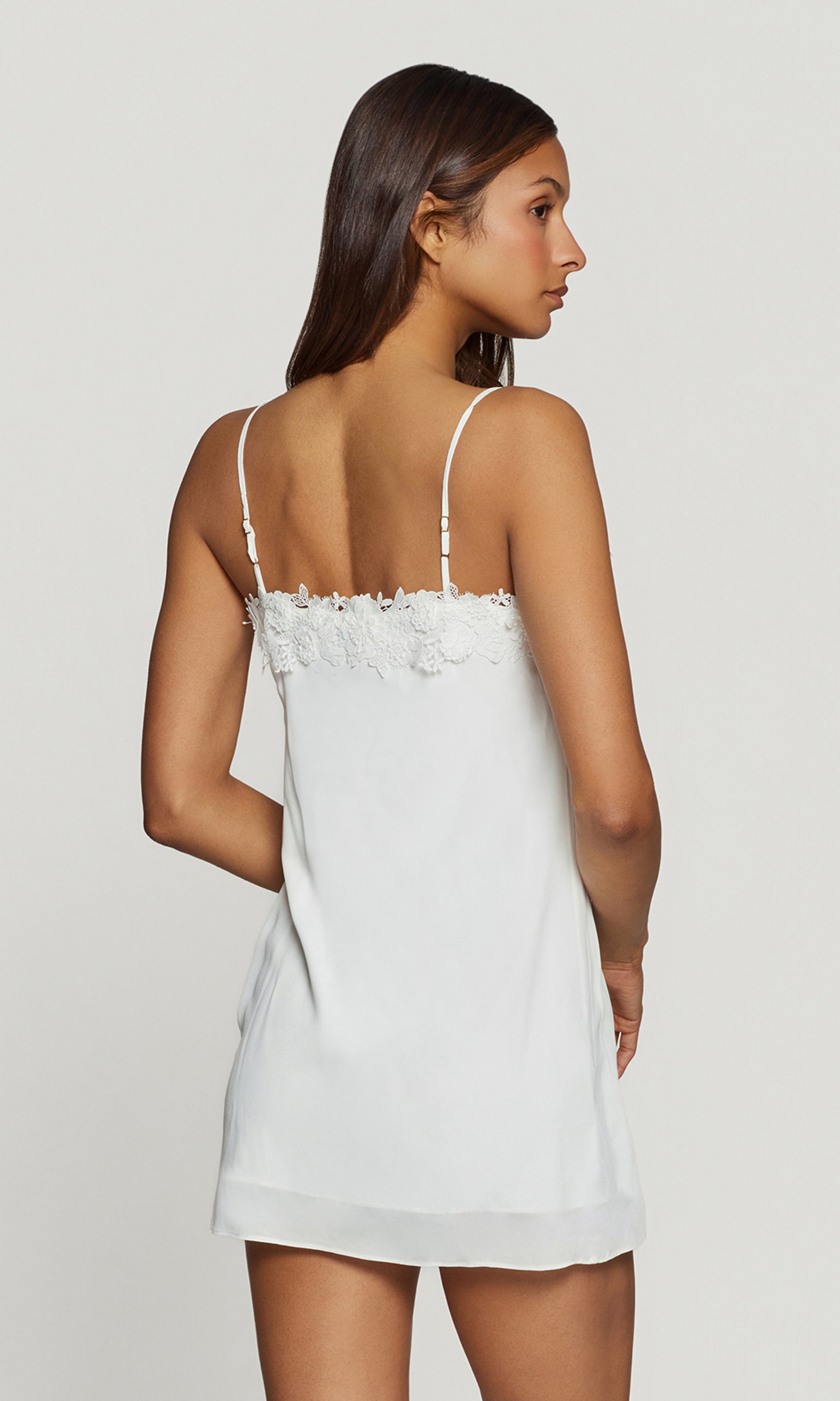 Back view of woman wearing a white lace-trimmed slip dress against a plain background
