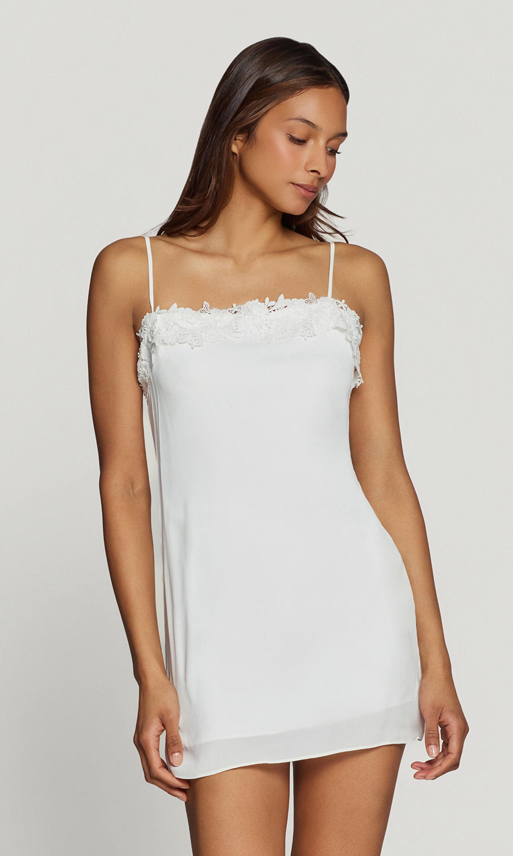 Side view of woman wearing a white slip dress with floral details on a plain background