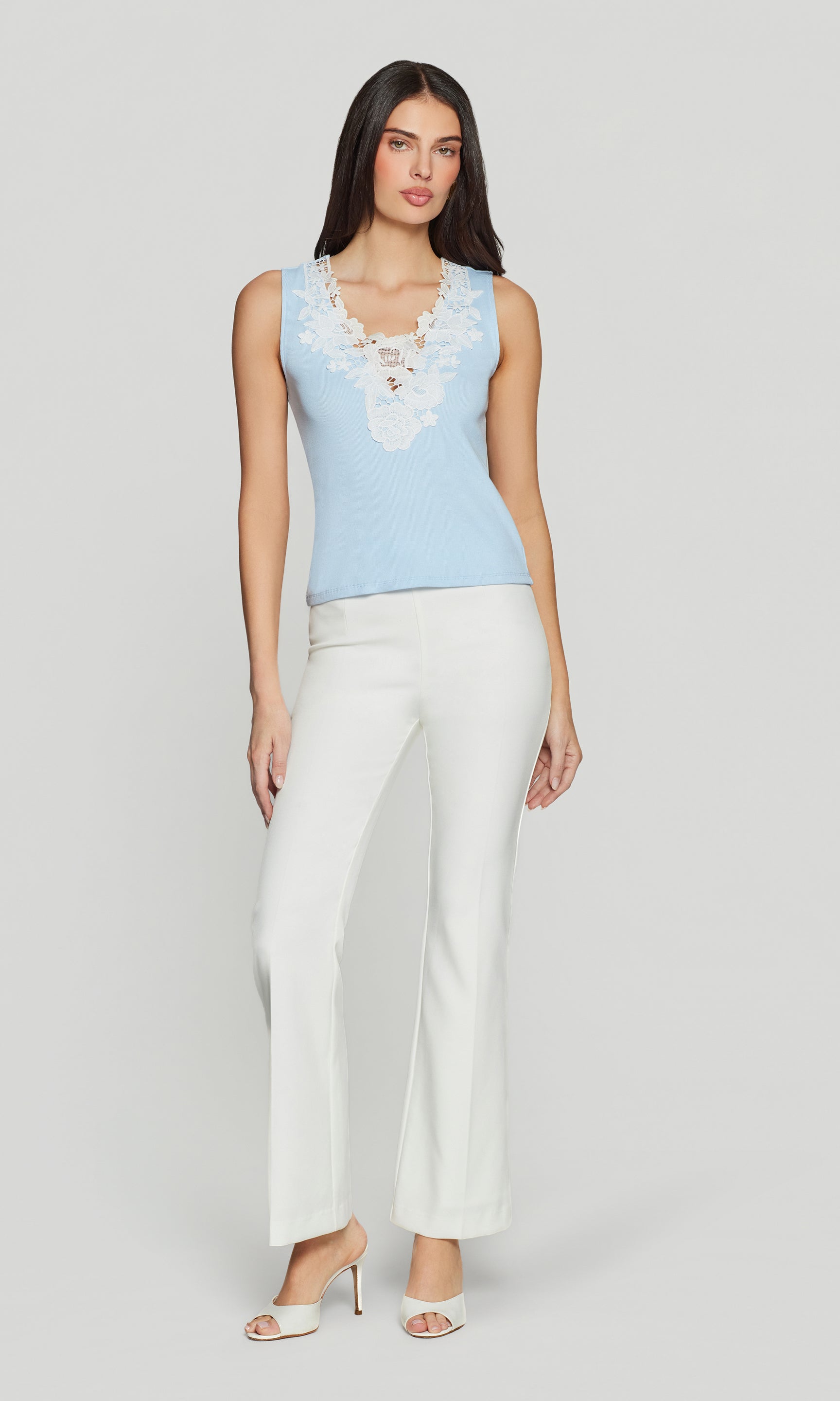 Woman wearing a light blue sleeveless top with lace details and white pants on a plain background
