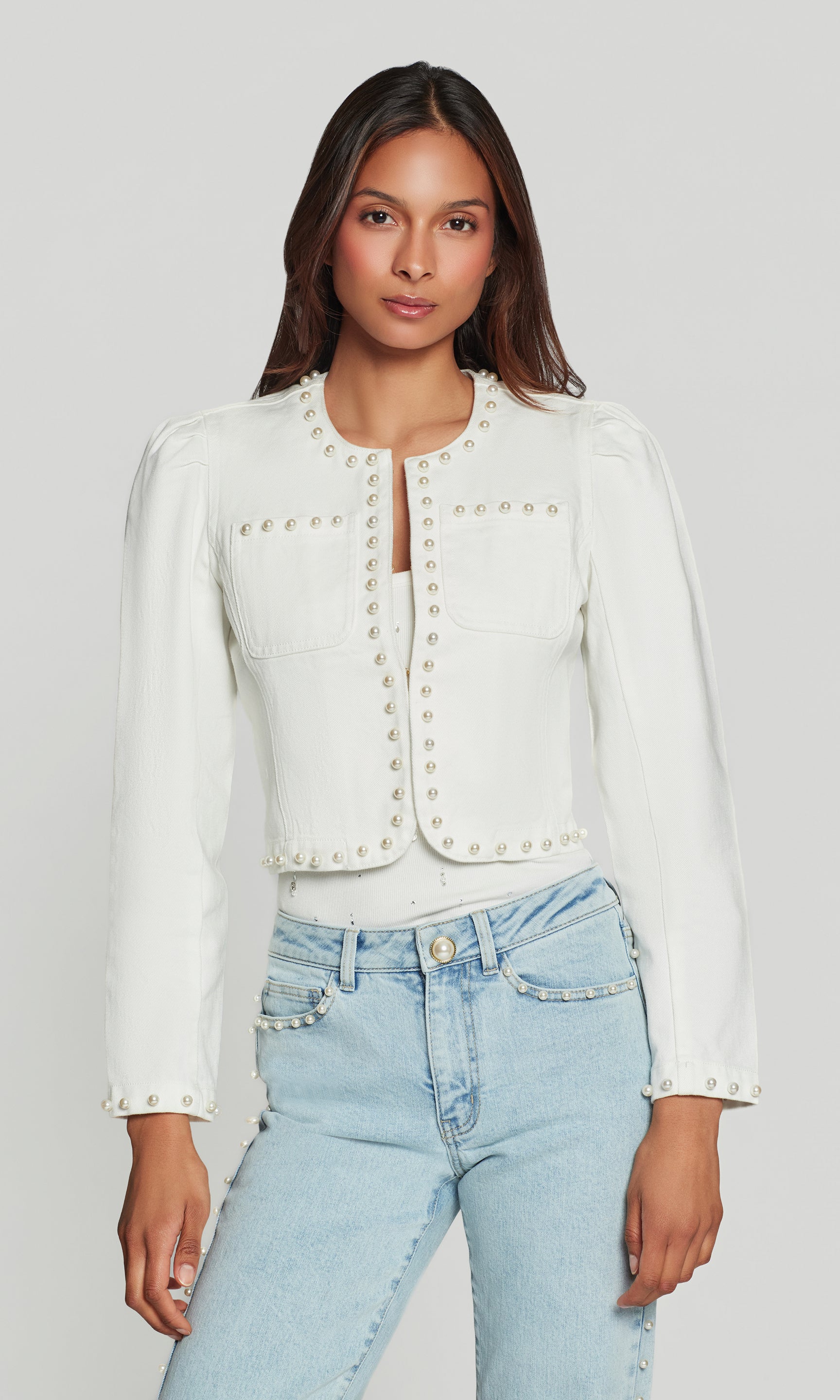 Woman wearing a white jacket with pearl buttons and light blue jeans on a plain background