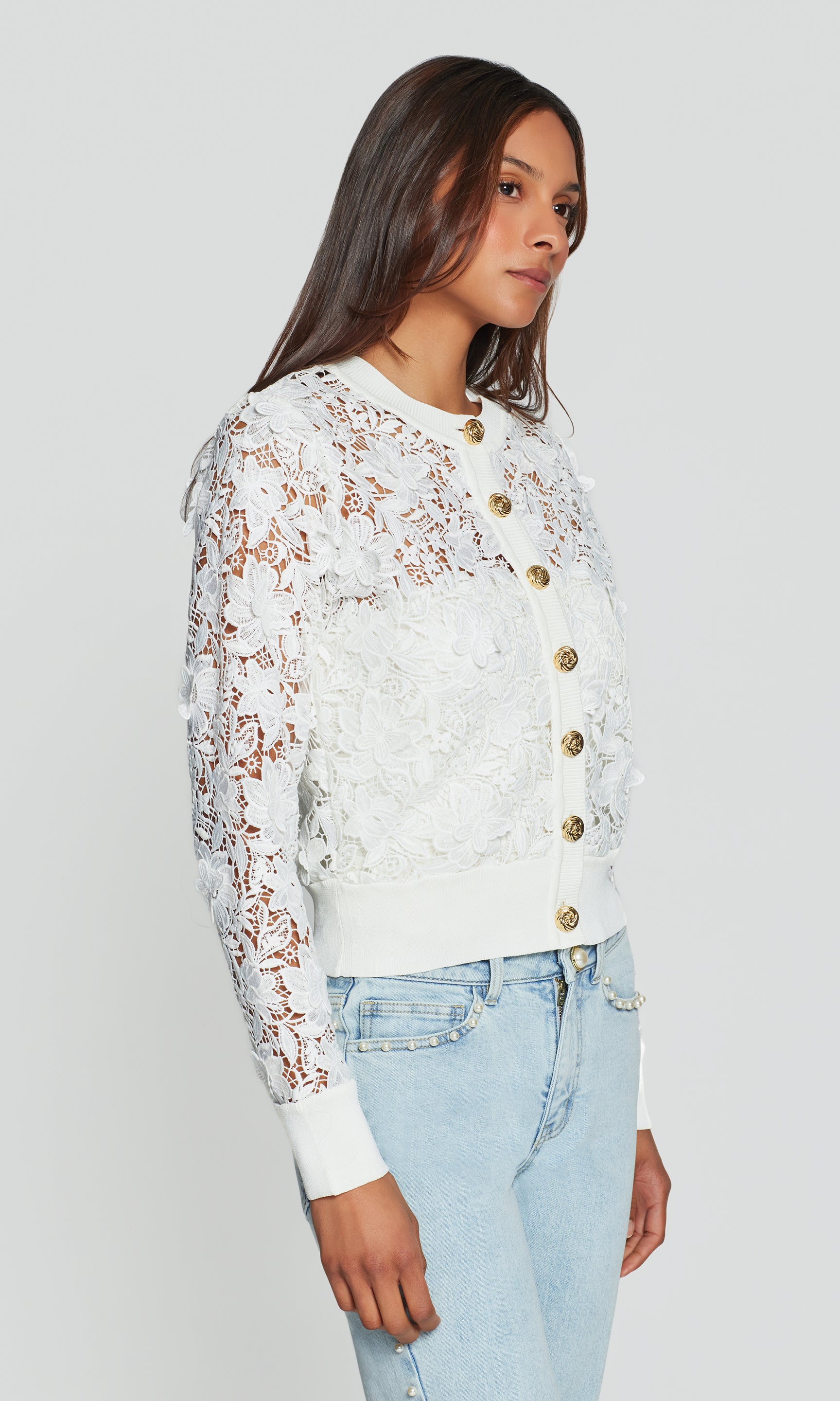 Side view of woman wearing a white lace cardigan with gold buttons on a plain background