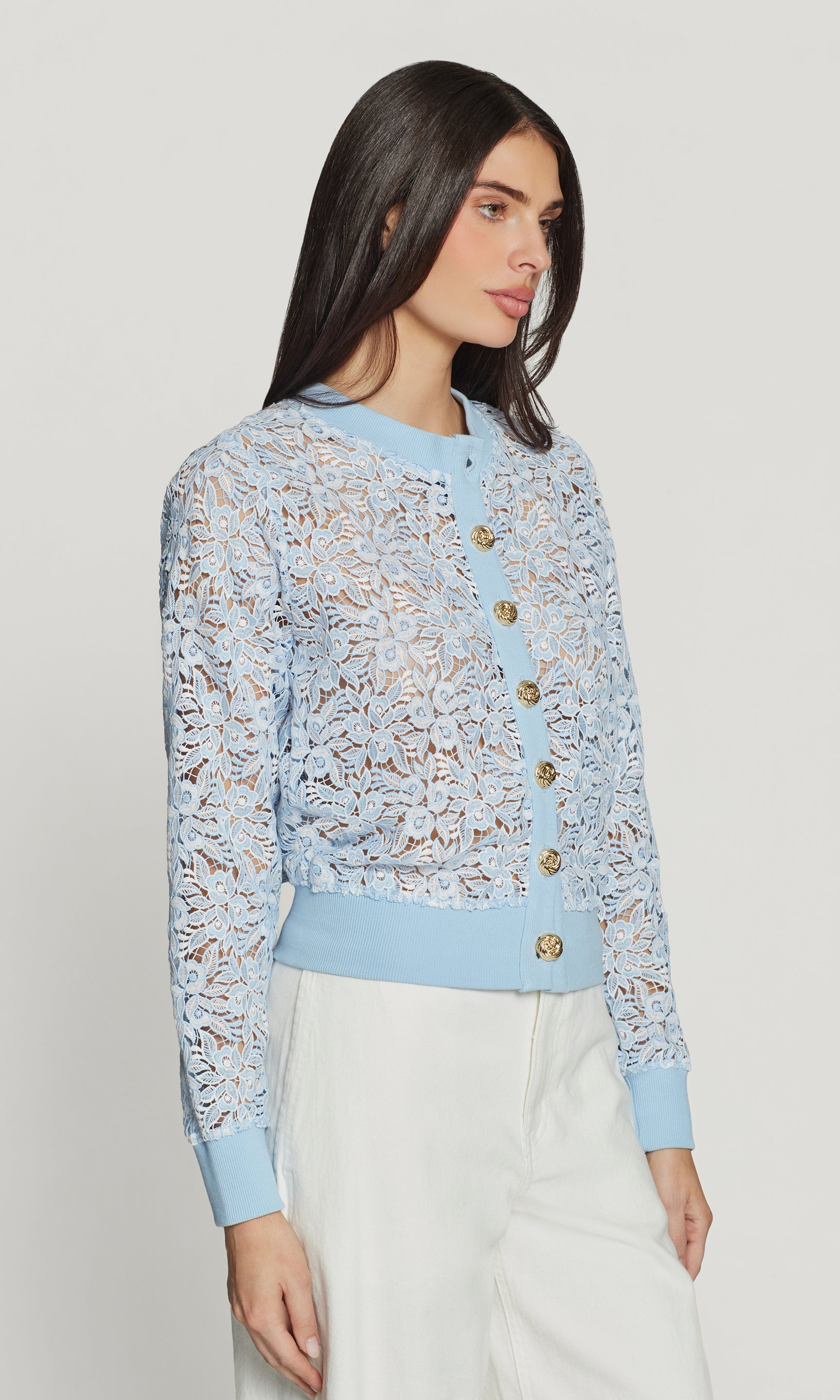 Side view of woman wearing a light blue lace cardigan with gold buttons on a plain background