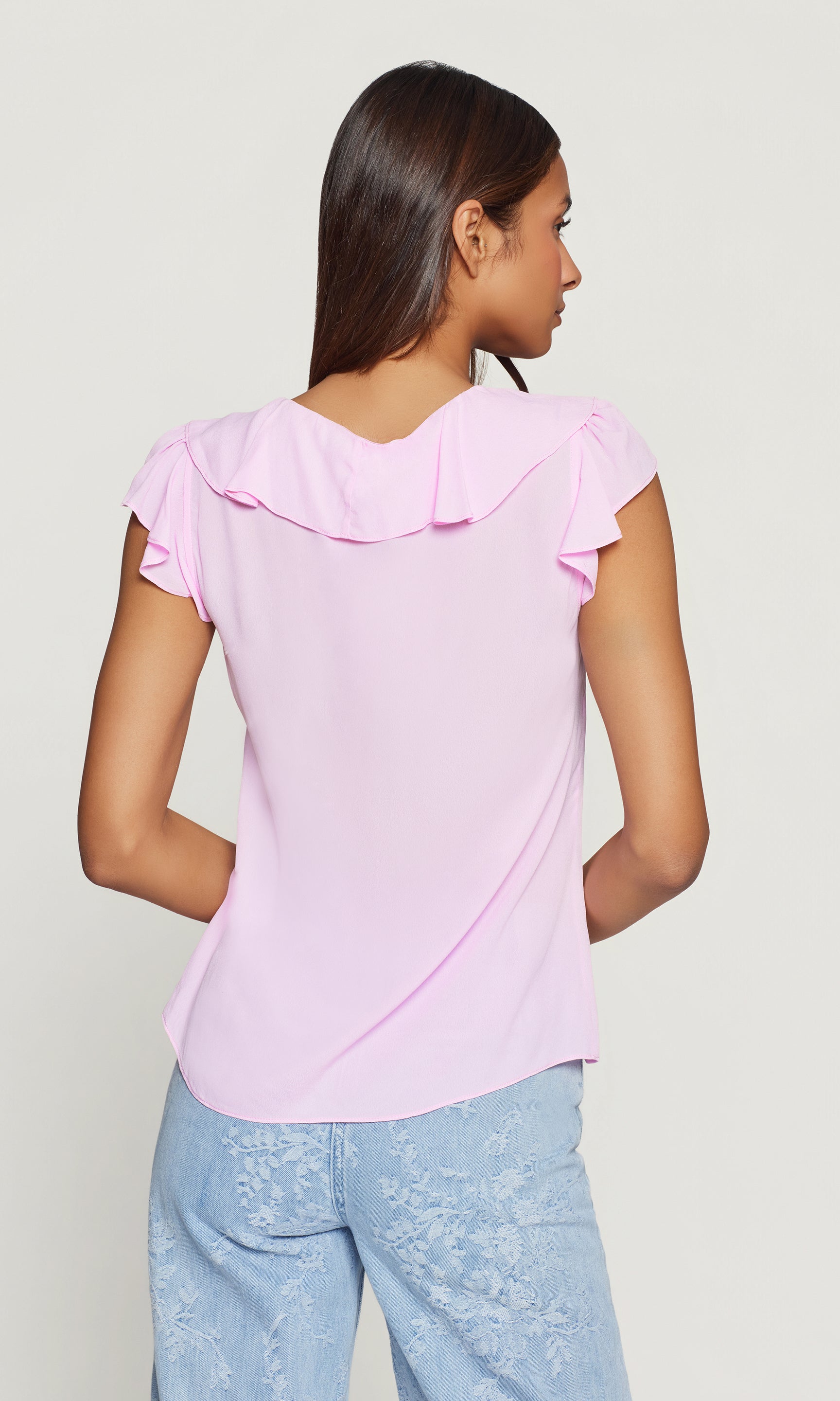 Back view of woman wearing a light pink blouse with ruffled details on a plain background