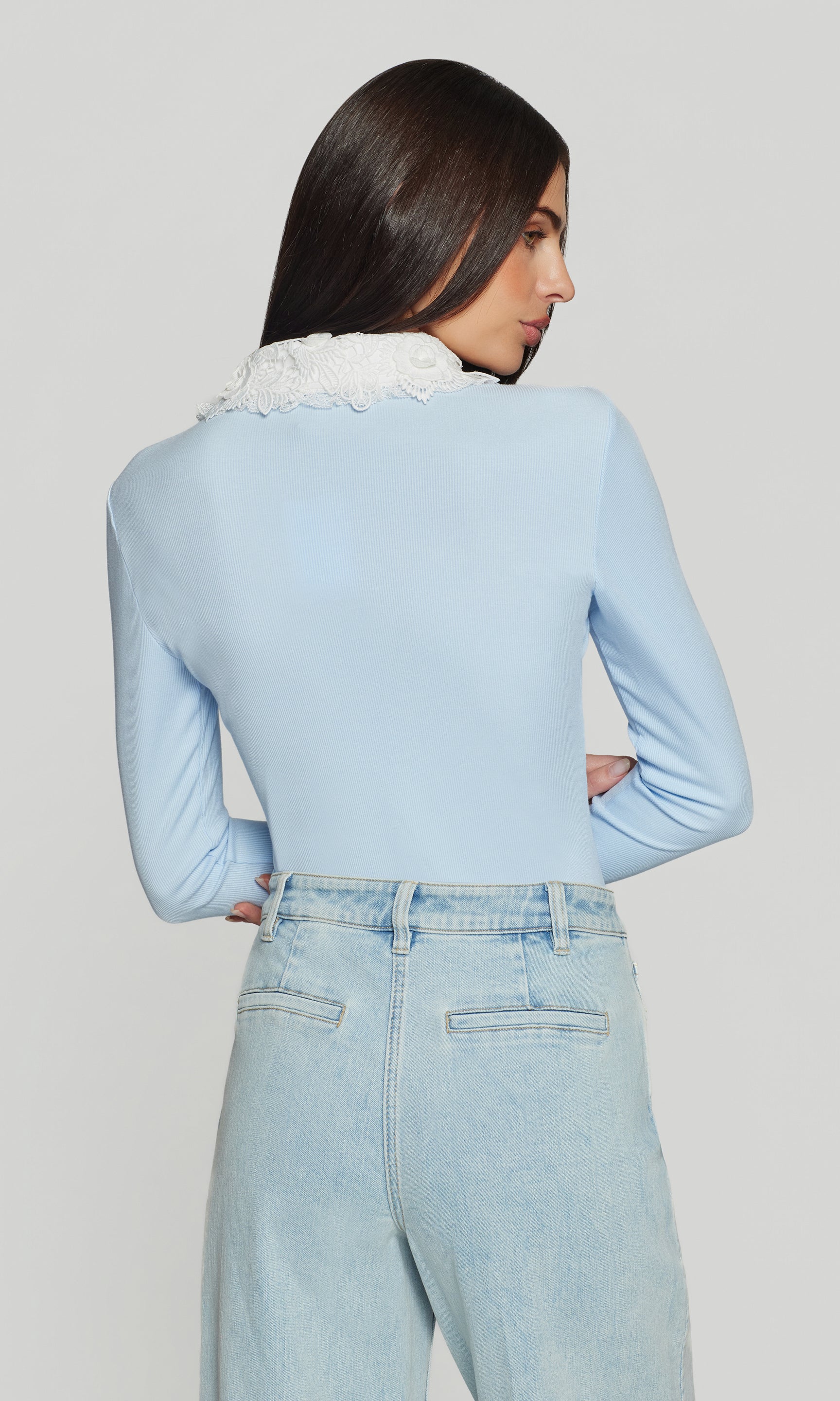 Back view of person wearing a light blue sweater with white collar and light blue jeans on a plain background