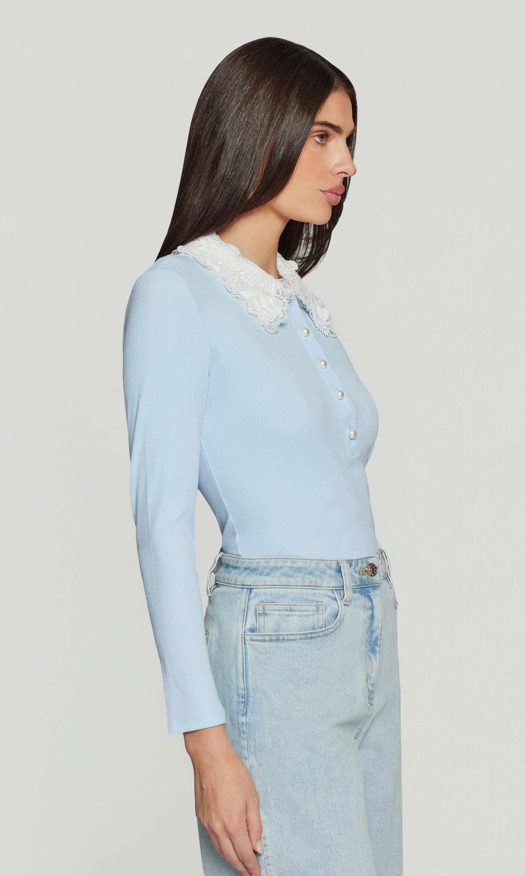Side view of woman wearing a light blue long-sleeve top with a white collar and light blue jeans on a gray background