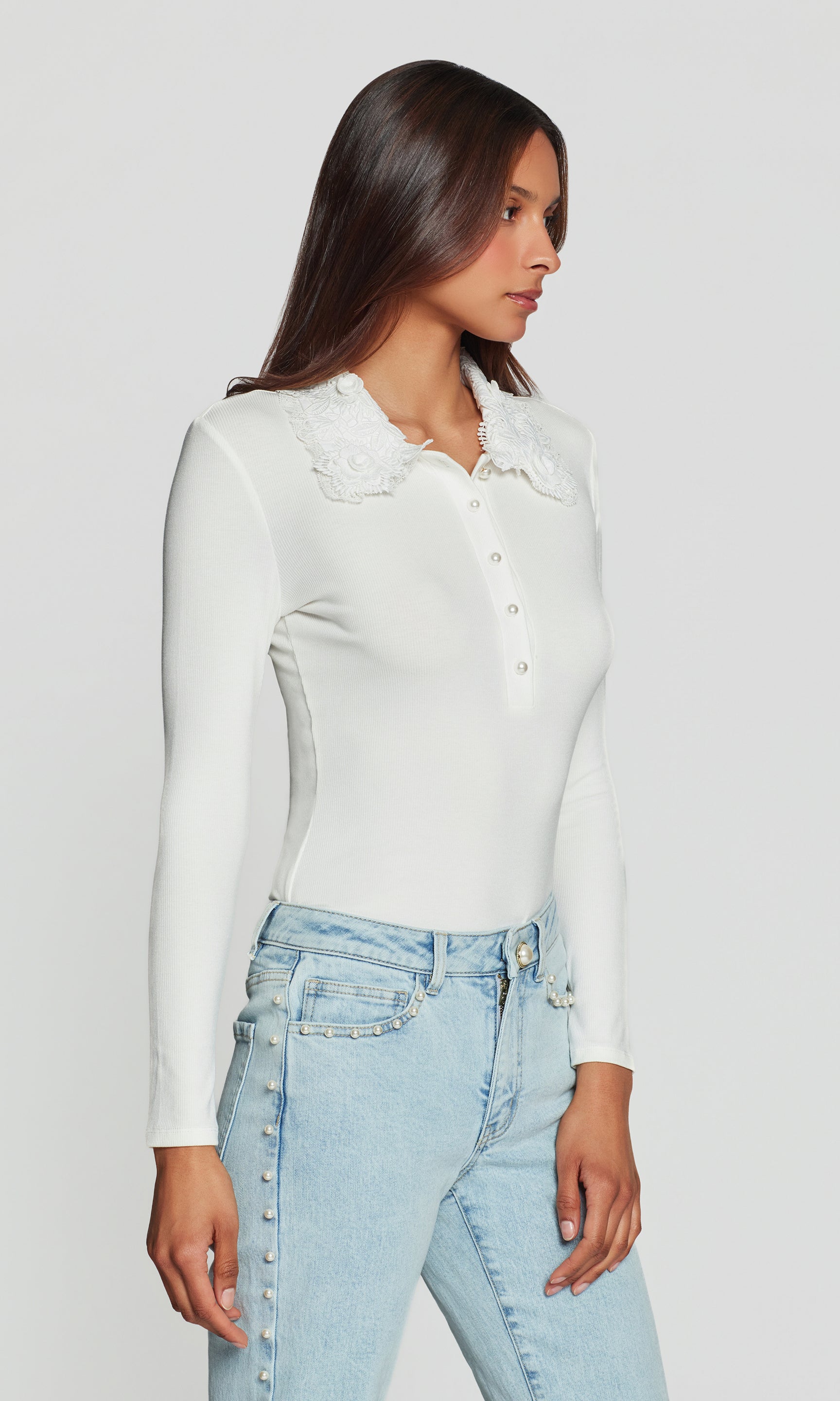 Side view of woman wearing a white long-sleeve shirt and light blue jeans on a plain background