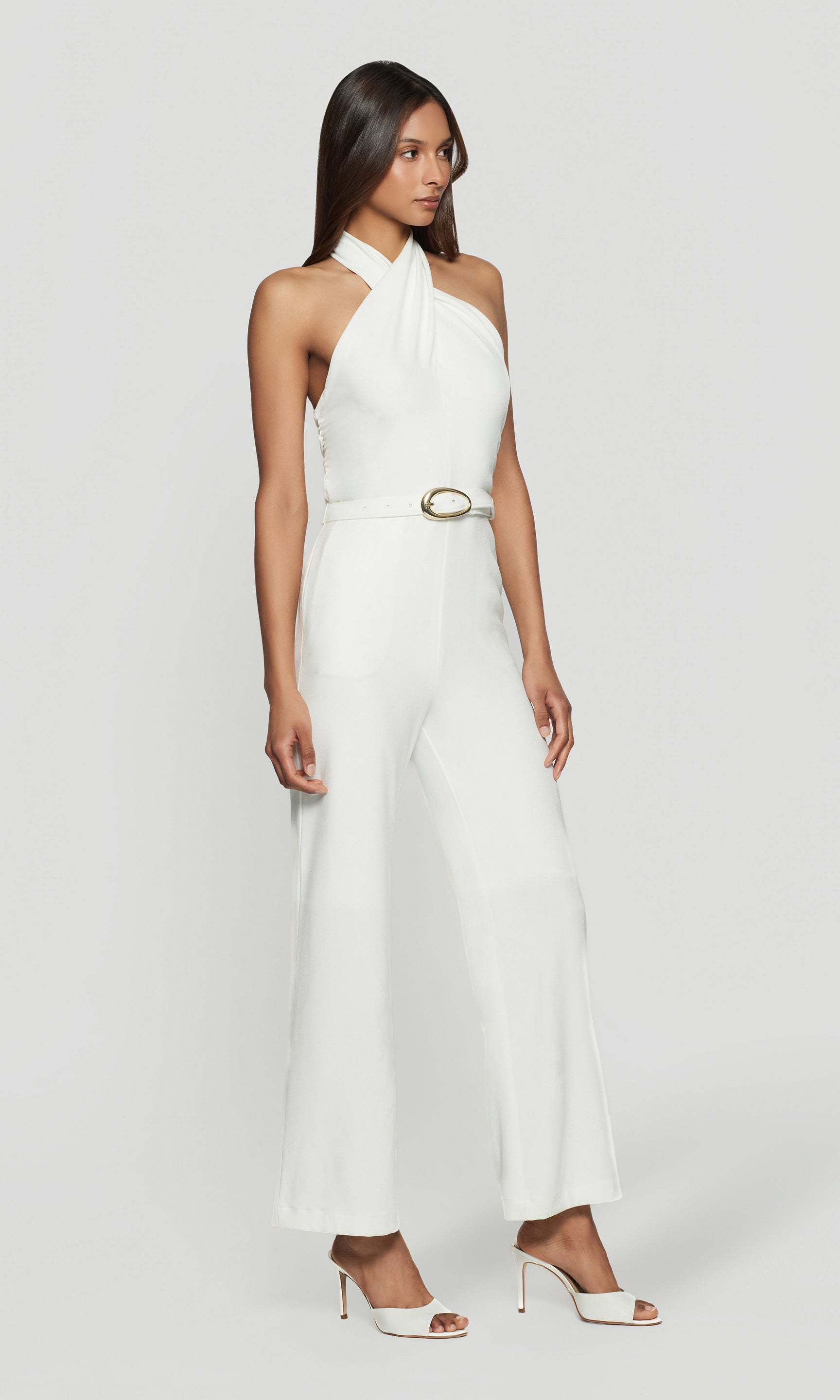 Side view of woman wearing a white halter neck jumpsuit on a plain background