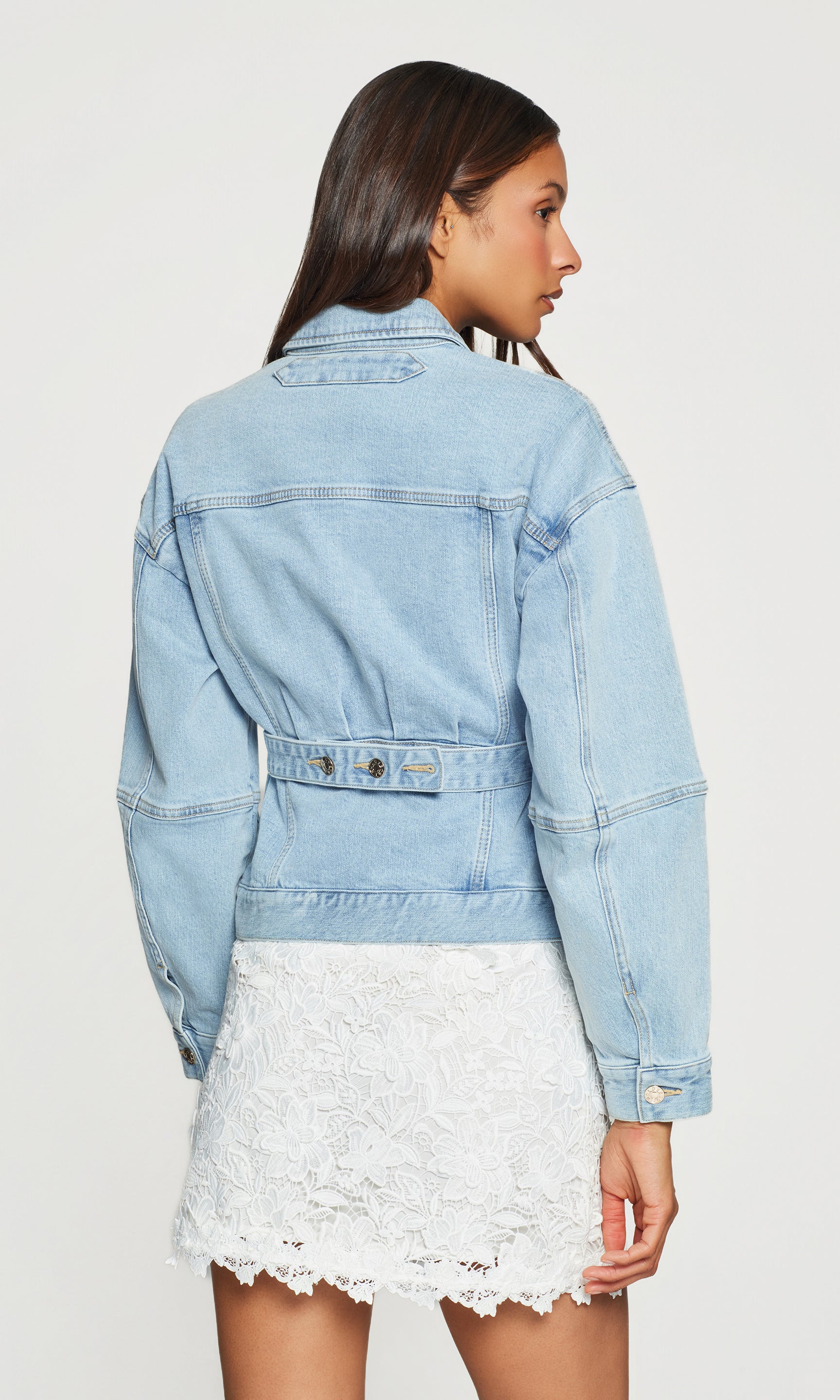 Back view of woman wearing a light blue denim jacket over a white lace skirt on a plain background
