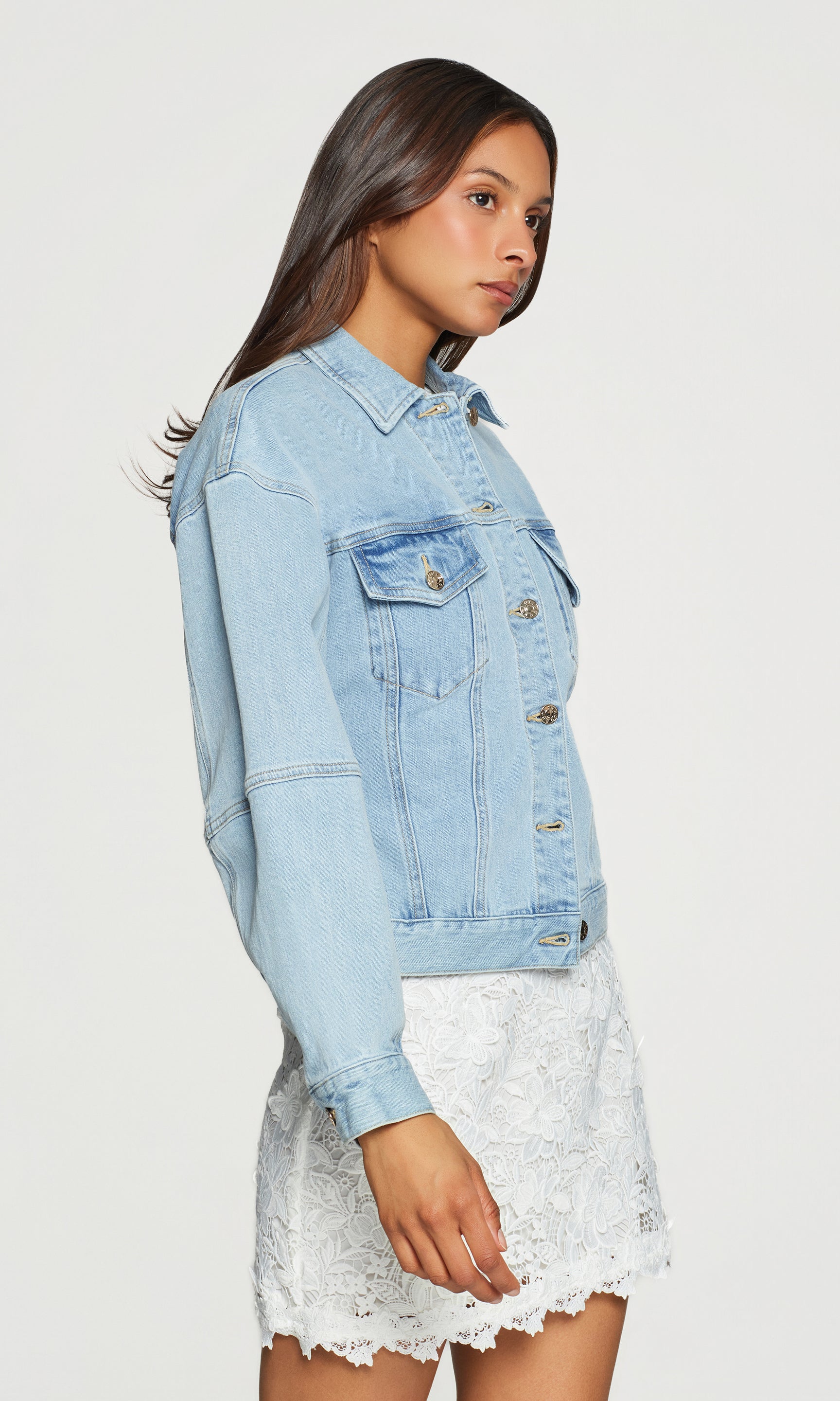 Side view of woman wearing a light blue denim jacket over a white lace dress on a plain background