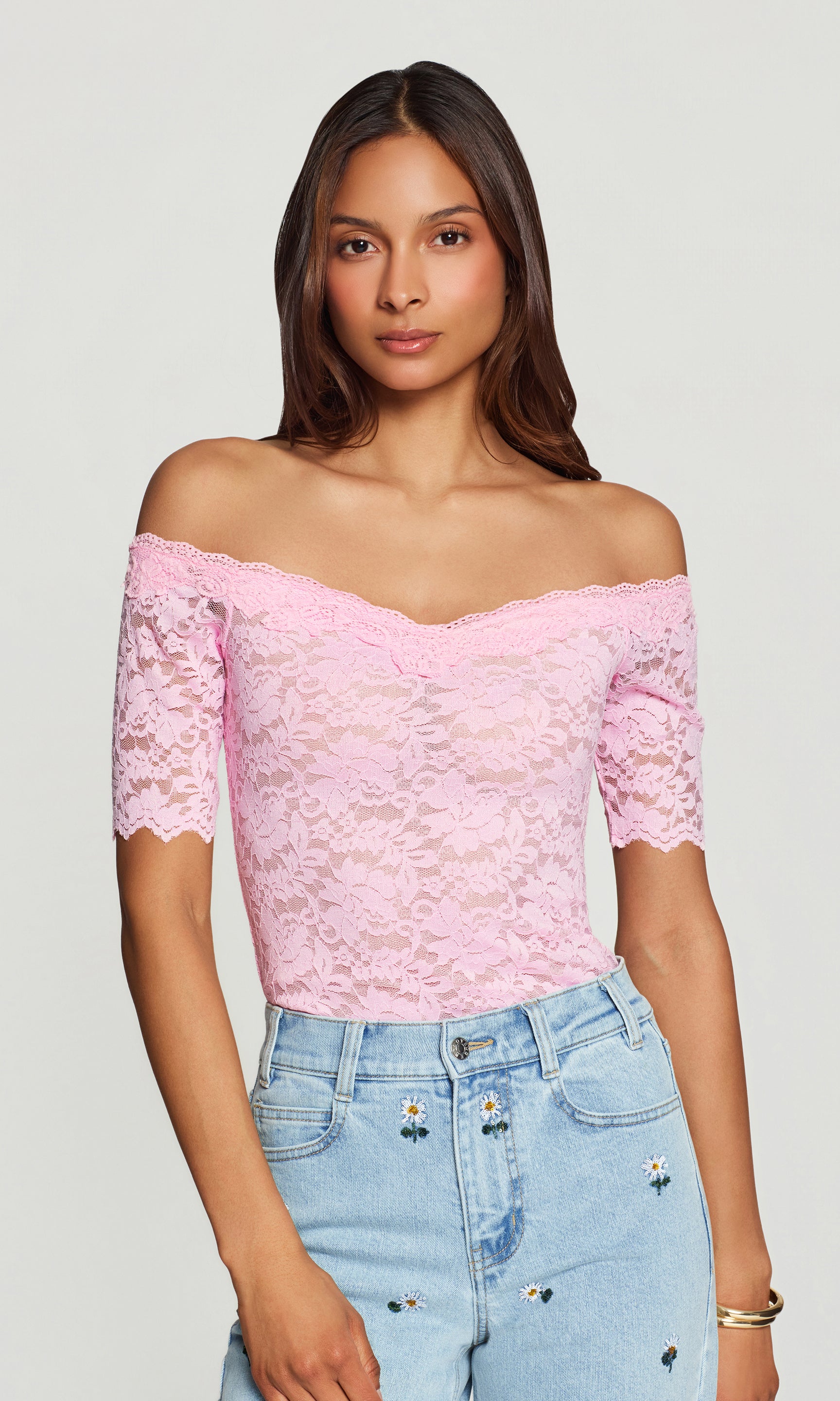 Woman wearing a pink lace off-shoulder top and blue jeans on a plain background