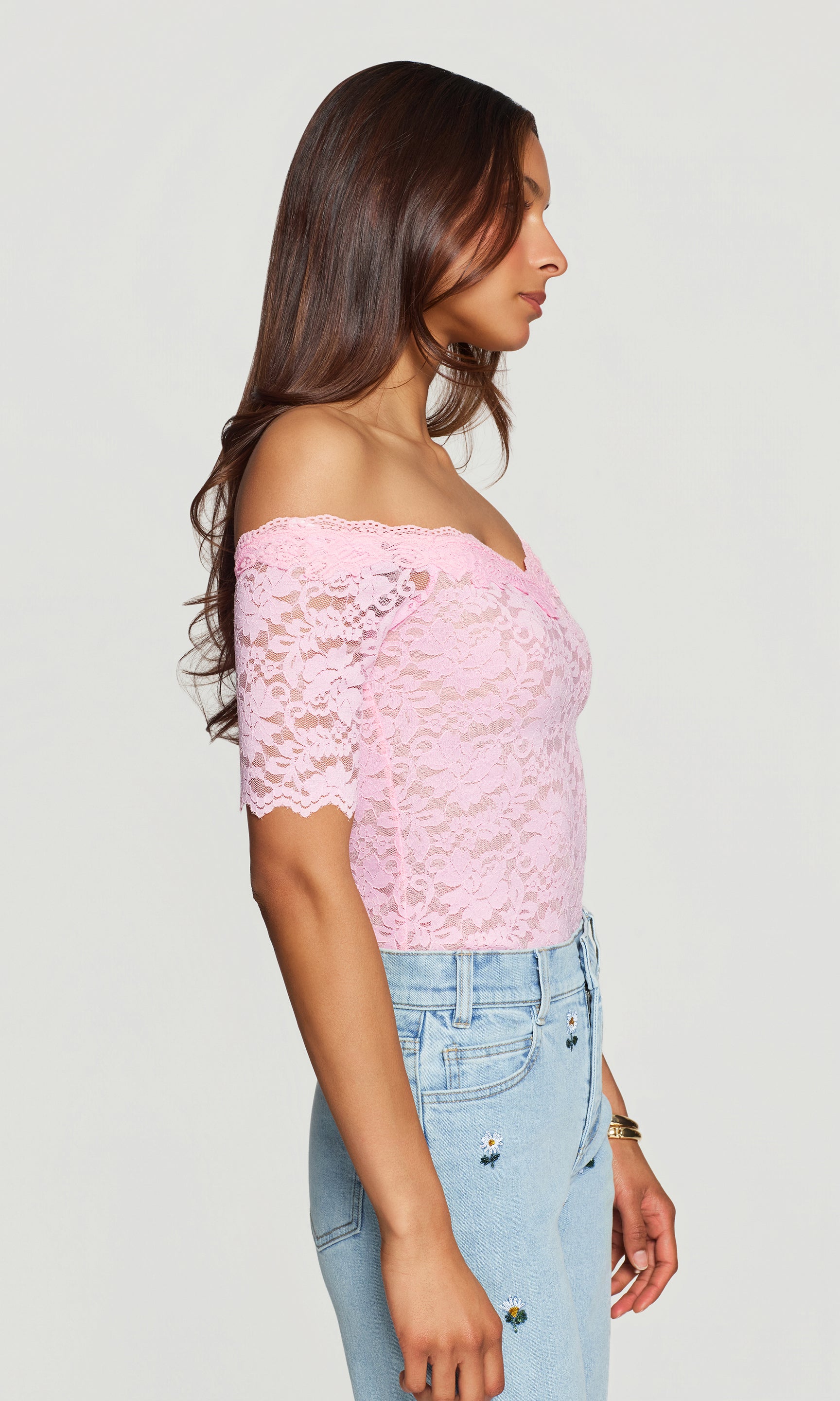 Side view of woman wearing a pink lace top and light blue jeans on a plain background