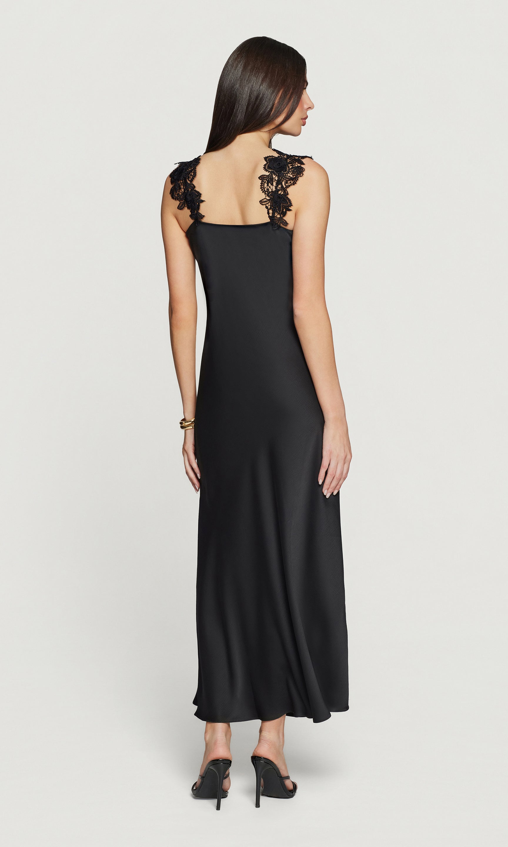 Back view of woman wearing a black sleeveless dress with floral lace details on a white background
