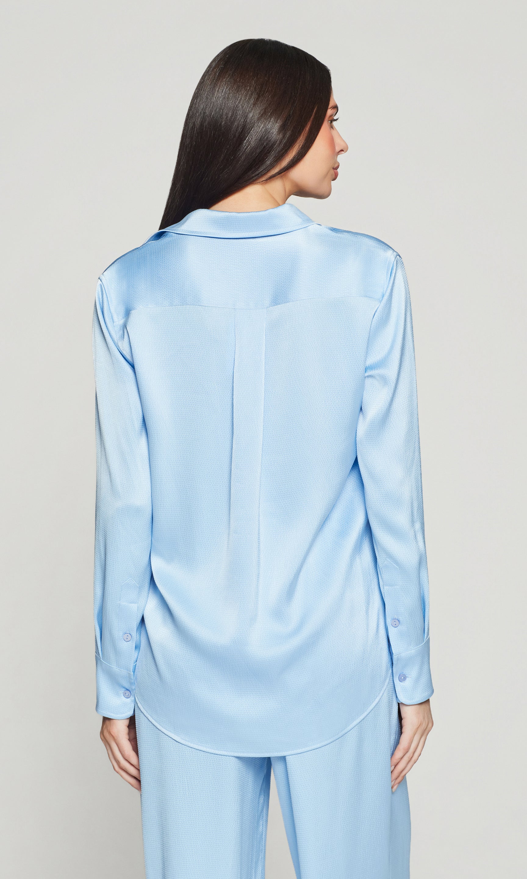 Back view of woman wearing a light blue satin shirt and pants on a plain background