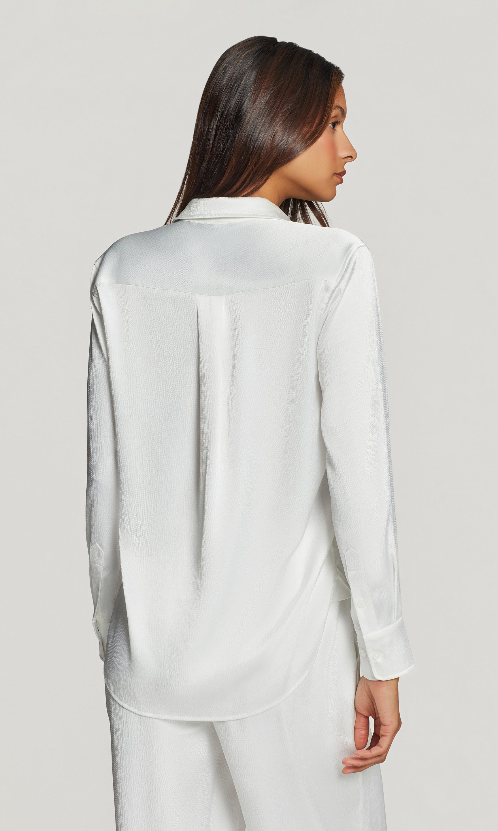 Back view of woman wearing a white satin shirt and pants on a light gray background