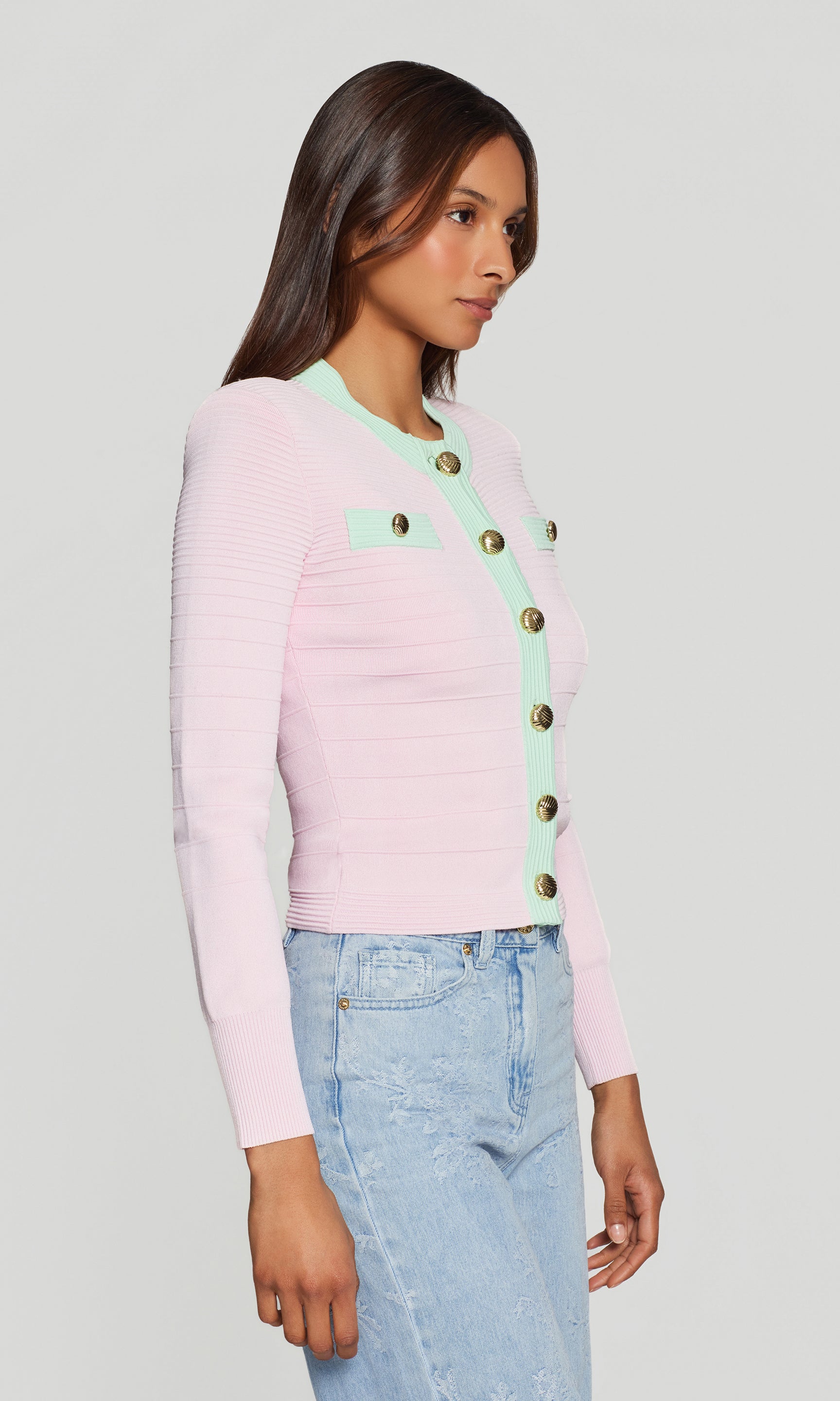 In a side view, the model wears the Lilla Bandage Cardigan in pink with denim, emphasizing the fitted profile and mint trim along the neckline and placket.