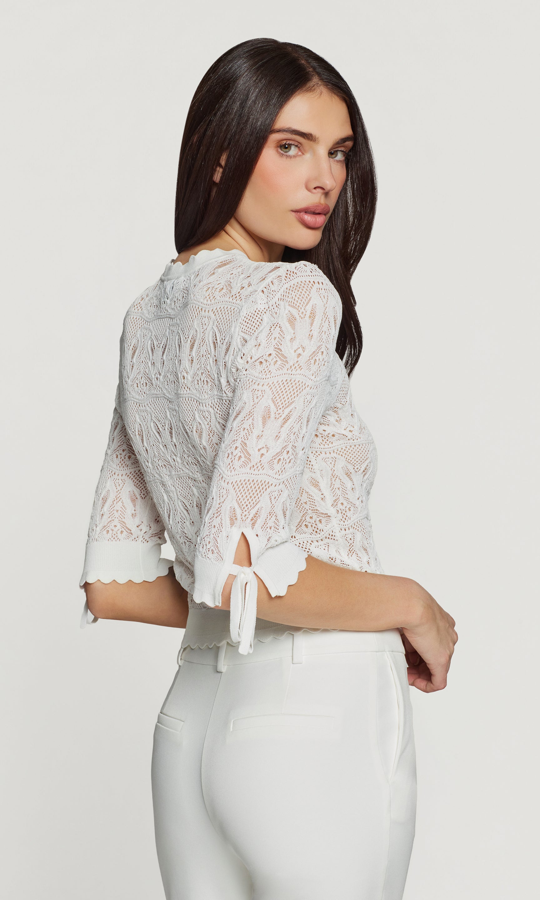 Back 3/4 view of woman wearing a white lace cardigan with butterfly knit detail on a plain background