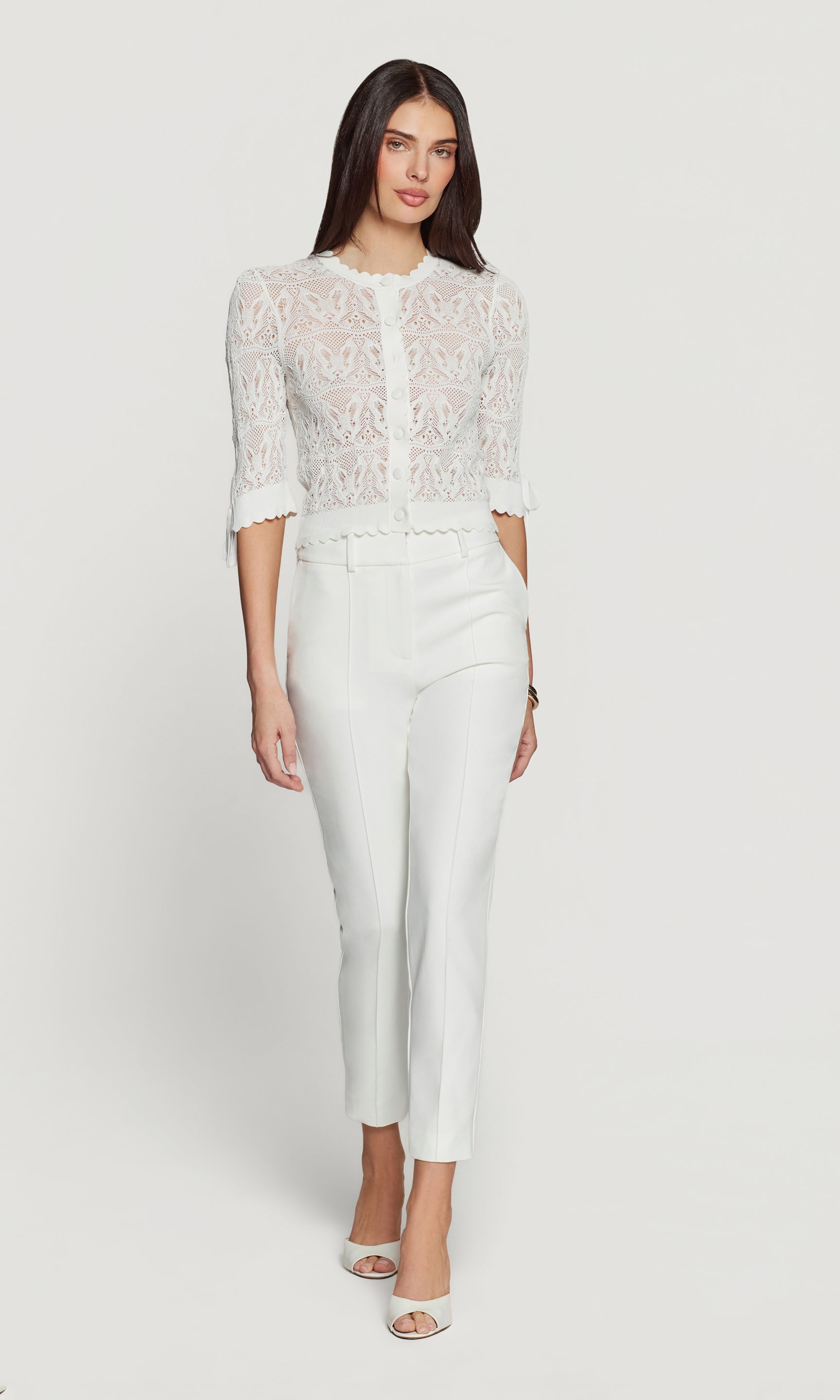 Woman wearing a white lace cardigan and white pants on a white background