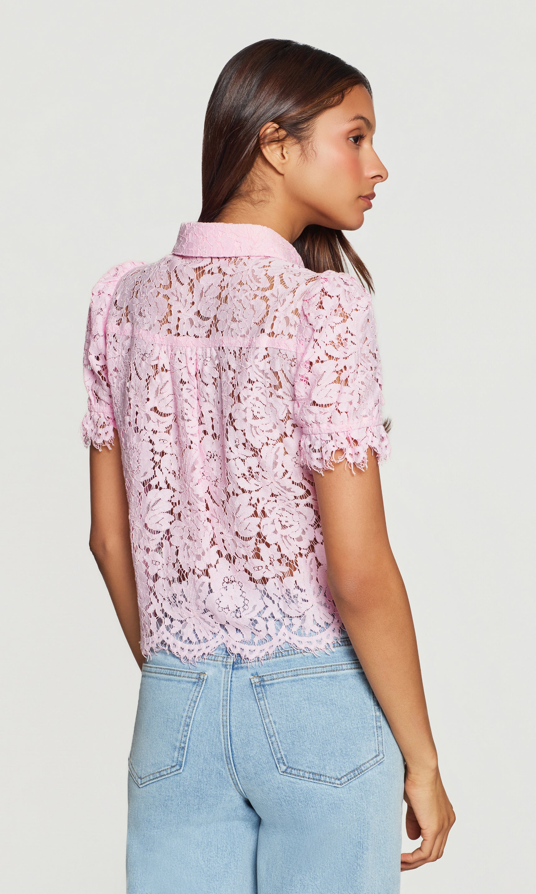 Back view of woman wearing a pink lace blouse and light blue jeans on a white background