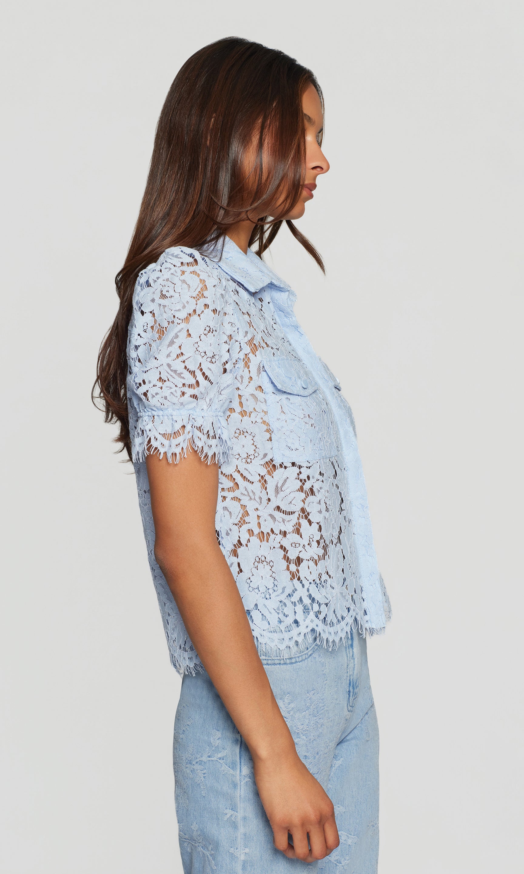 Side view of woman wearing a light blue lace top and jeans against a plain background