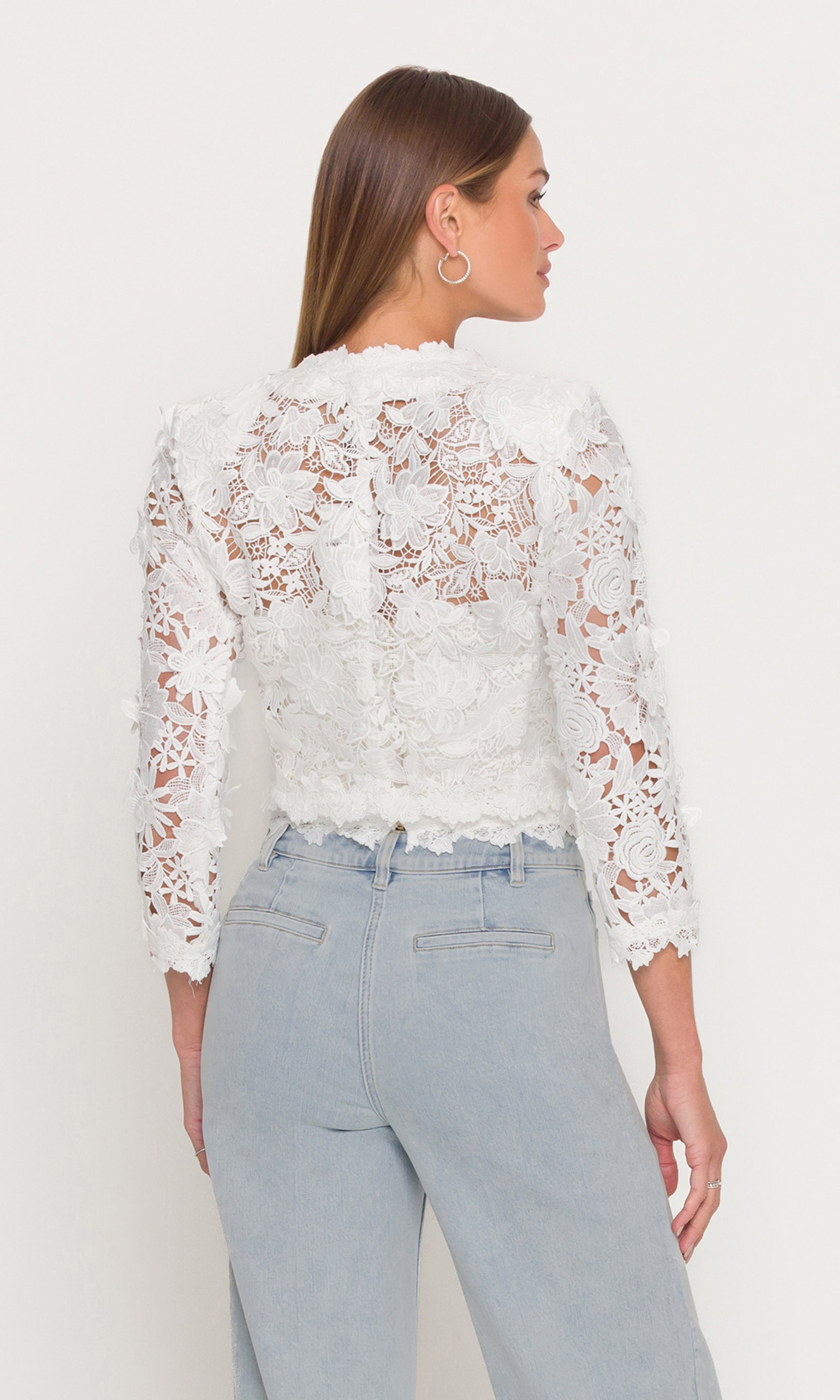 Back view of woman wearing a white lace jacket and light blue jeans on a plain background