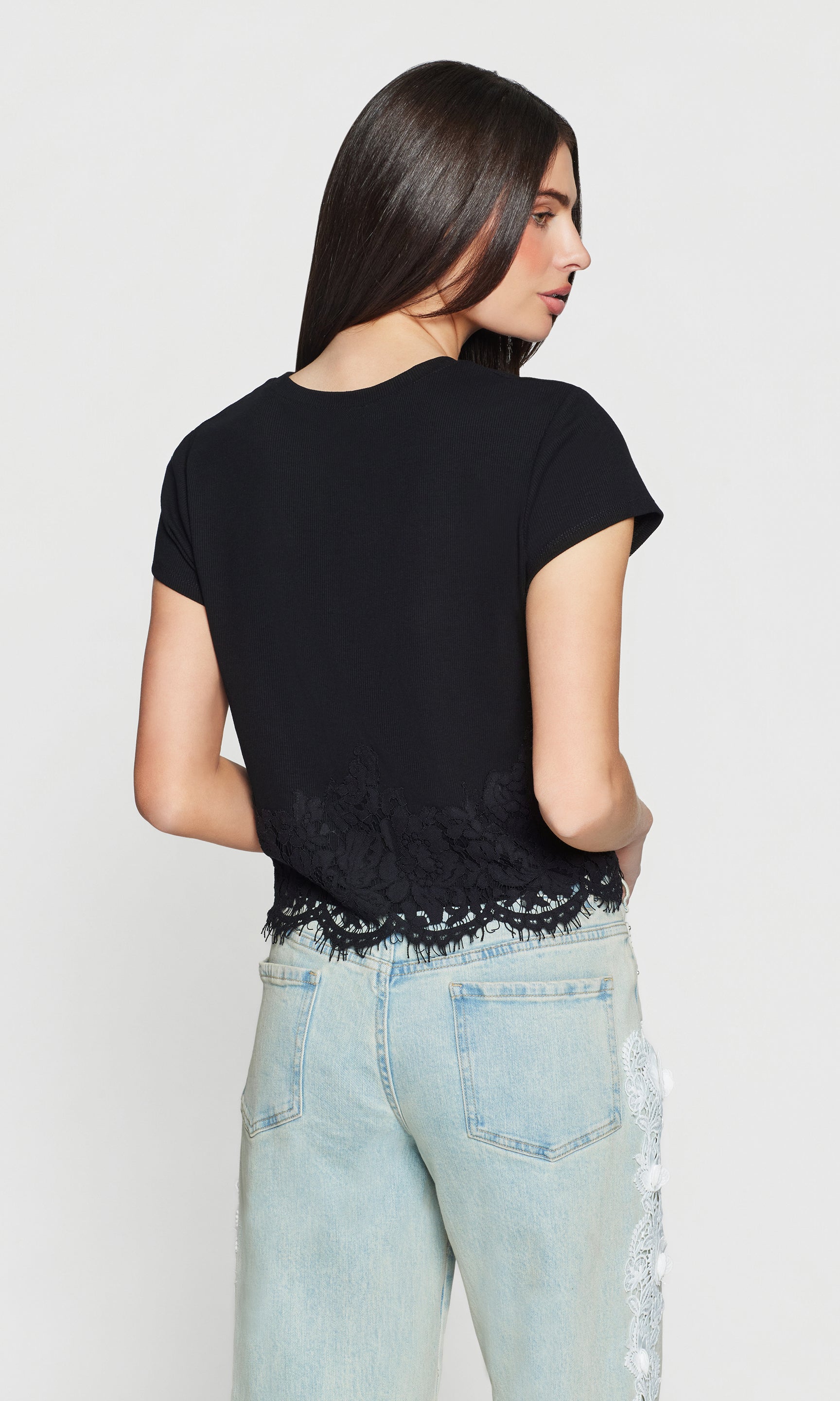 Back view of woman wearing a black top with lace trim and light blue jeans on a white background
