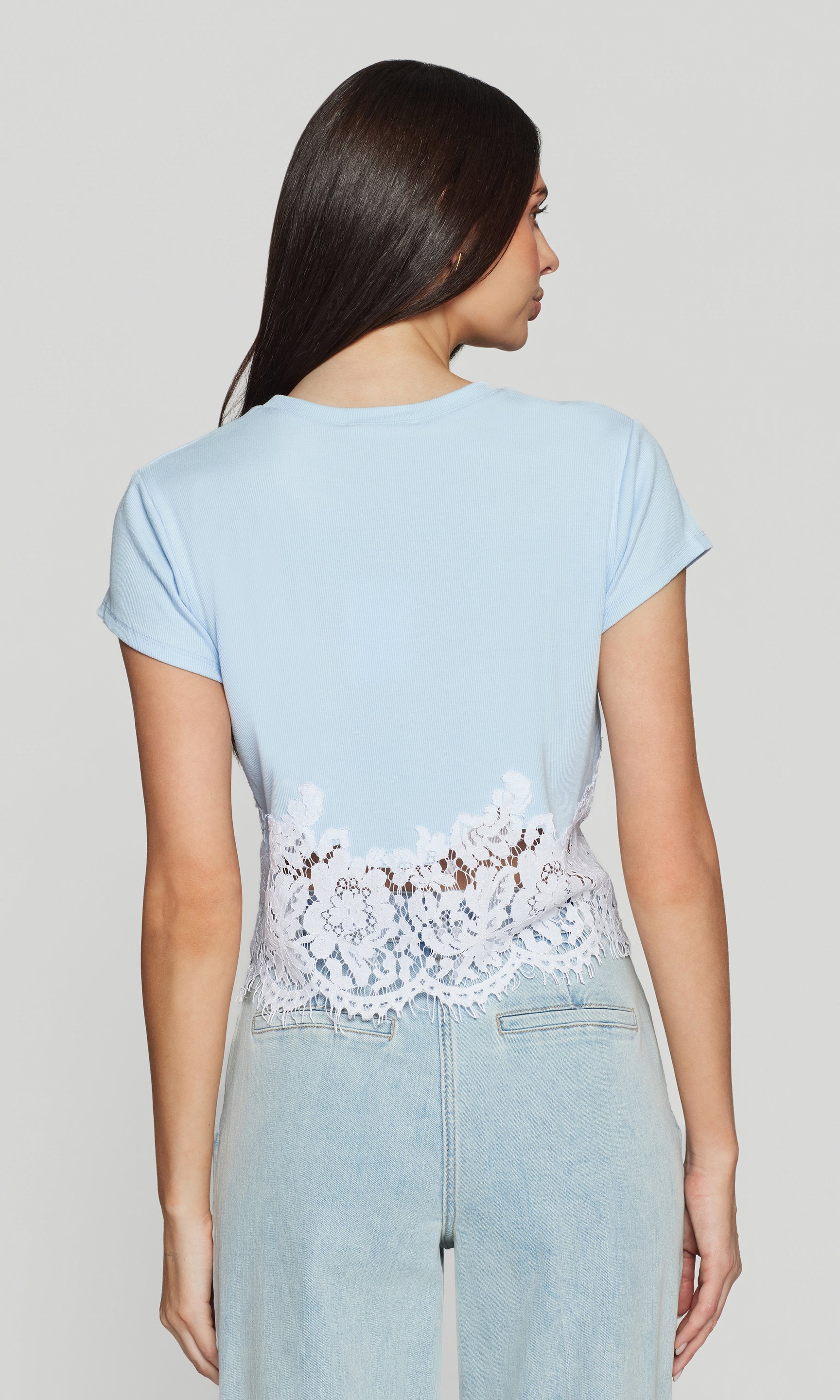 Back view of woman wearing a light blue top with lace detailing and light blue jeans on a plain background