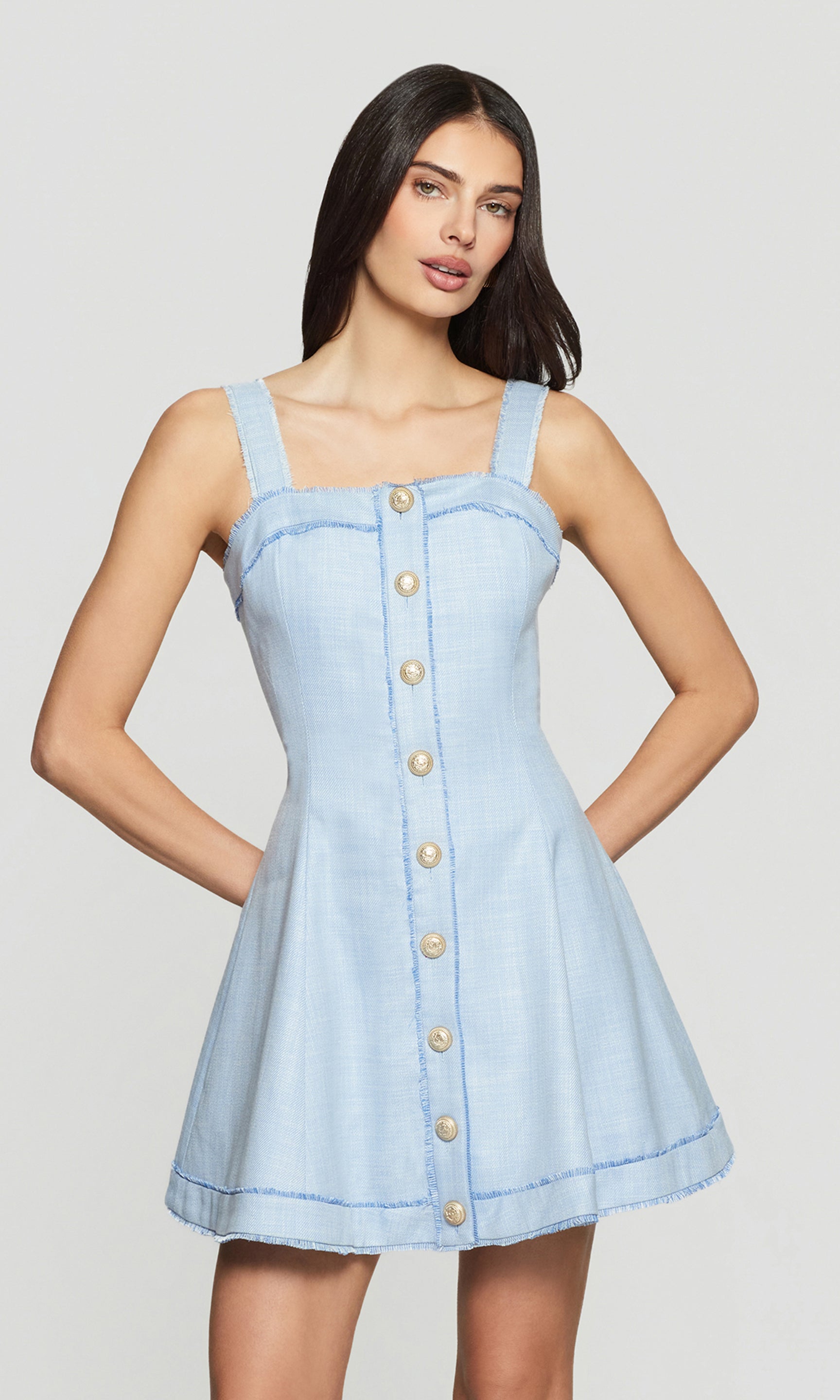 Model wears a chambray blue button-front mini dress in a full body front view, with wide straps and a flared skirt.
