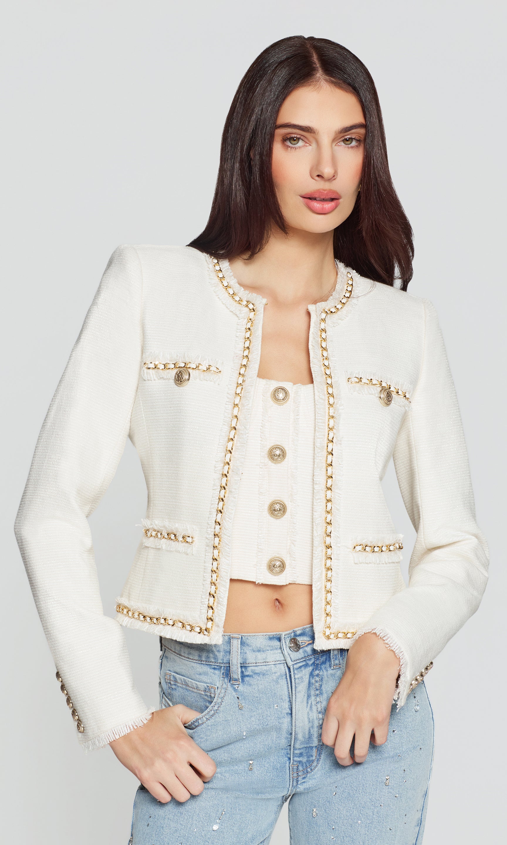 Woman wearing a white jacket with gold accents and blue jeans on a light gray background