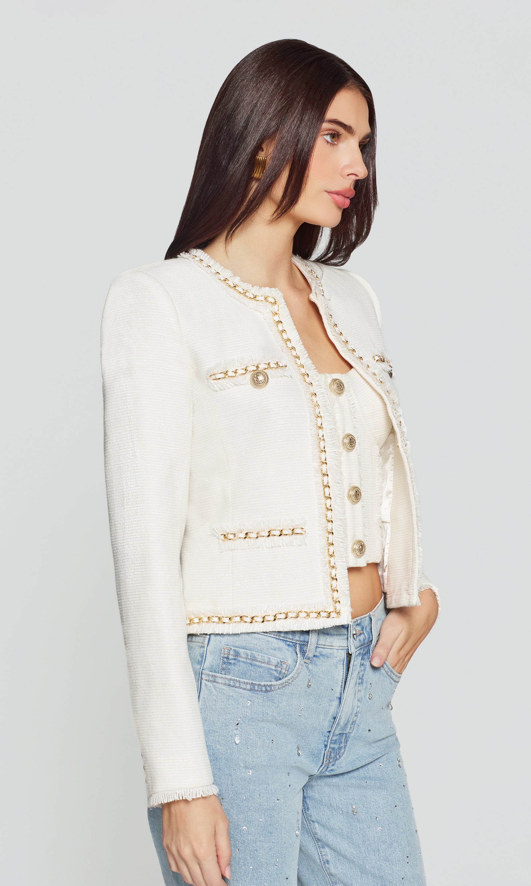 Side view of woman wearing a white jacket with gold buttons and light blue jeans on a plain background