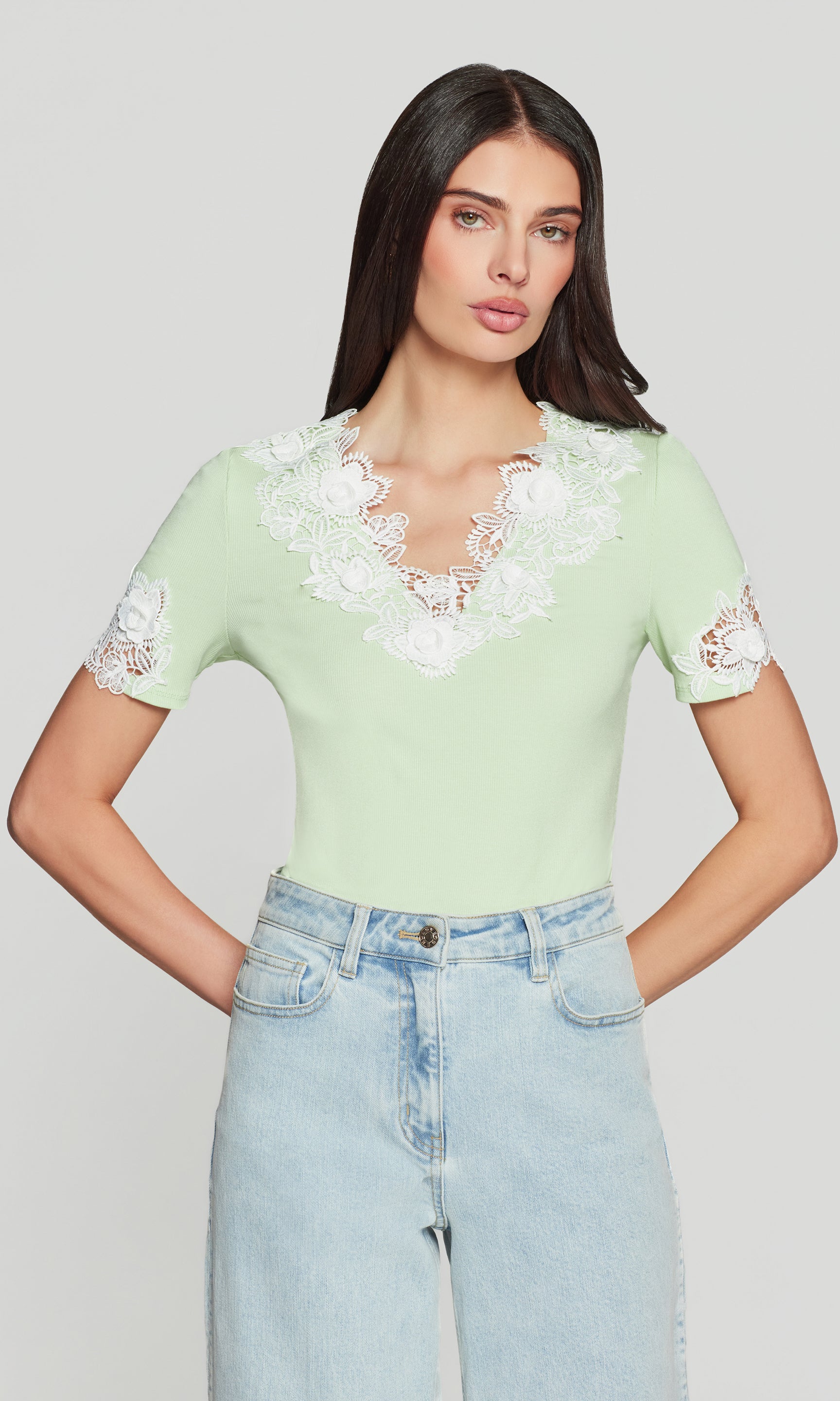 Woman wearing a light green lace-trimmed top and blue jeans on a plain background