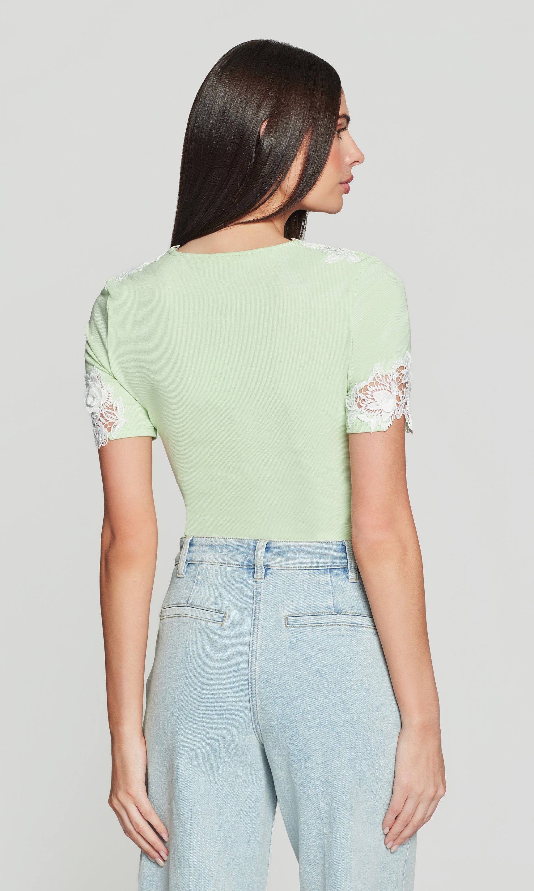 Back view of woman wearing a light green top with lace sleeves and light blue jeans on a plain background