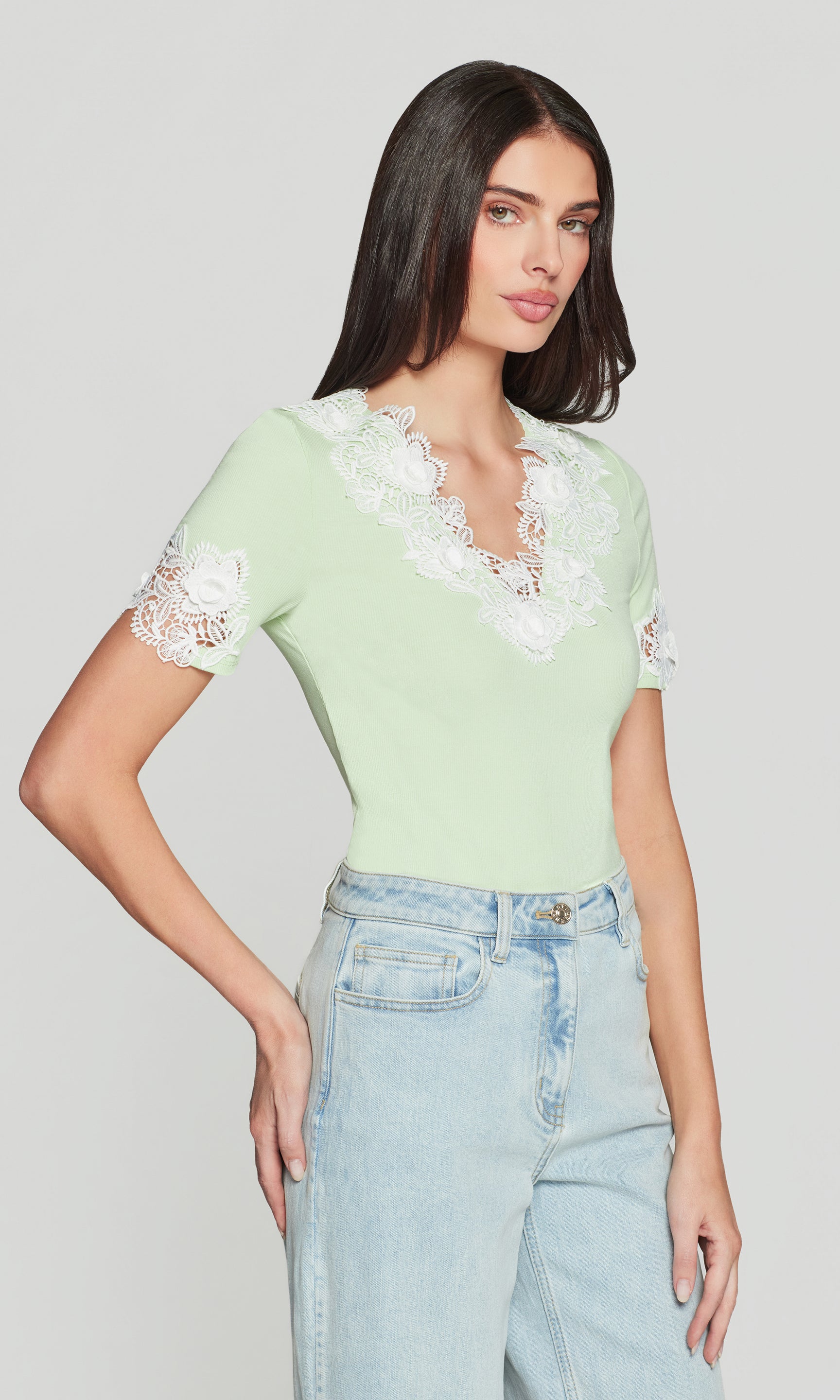 Side view of woman wearing a light green top with lace details and high-waisted jeans on a plain background
