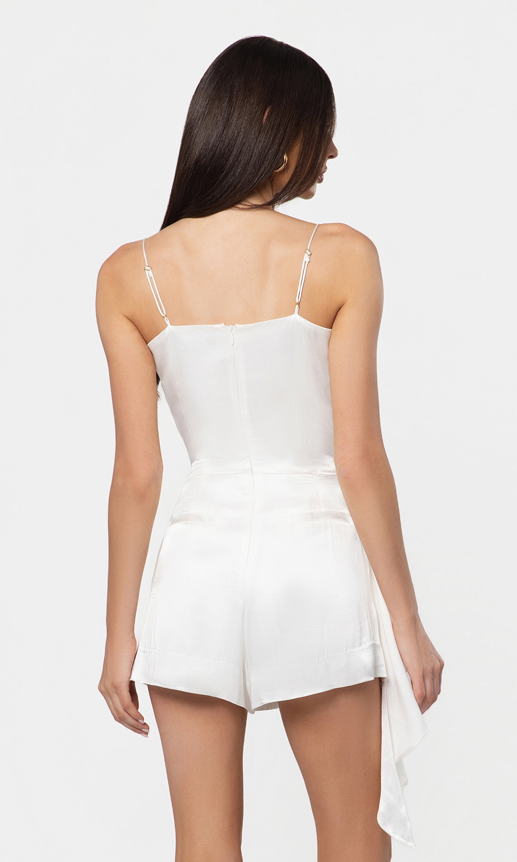 Back view of woman wearing a white romper with thin straps on a plain background