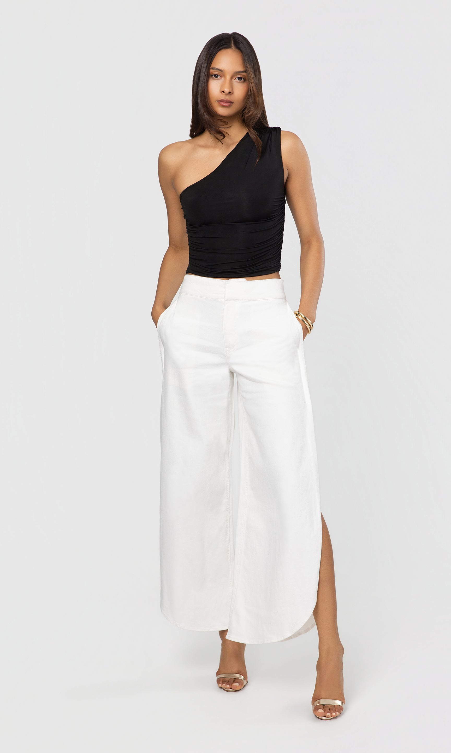 Woman wearing a black one-shoulder top and white pants with a high slit on a white background