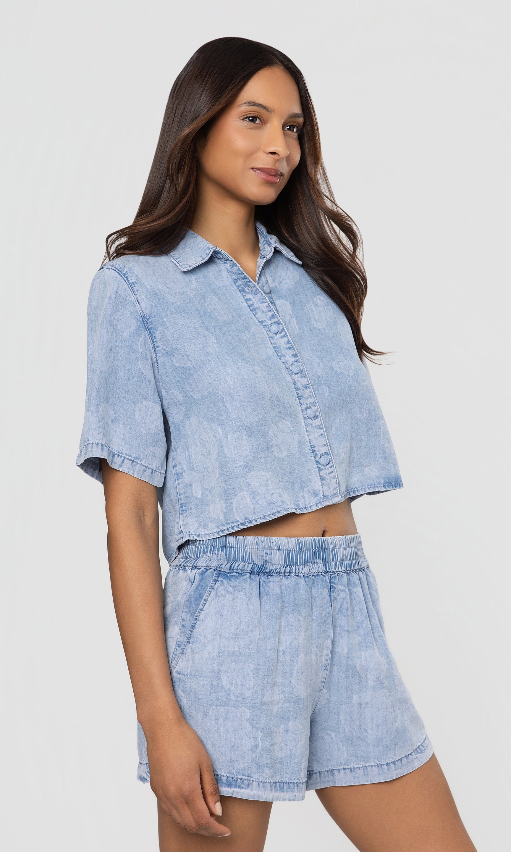 Side view of woman wearing a light blue denim shirt and shorts outfit on a white background