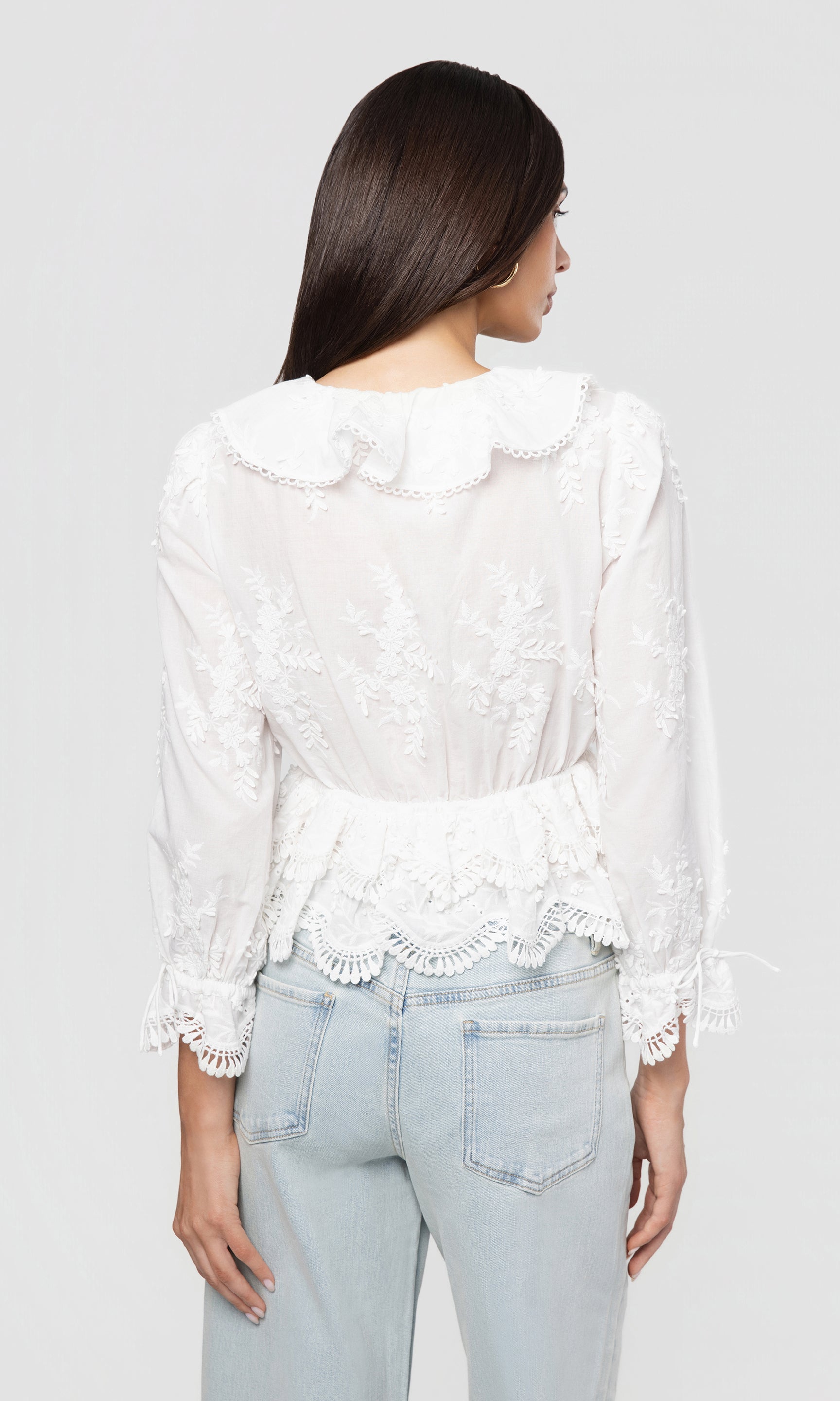 Back view of woman wearing a white lace blouse and light blue jeans on a plain background