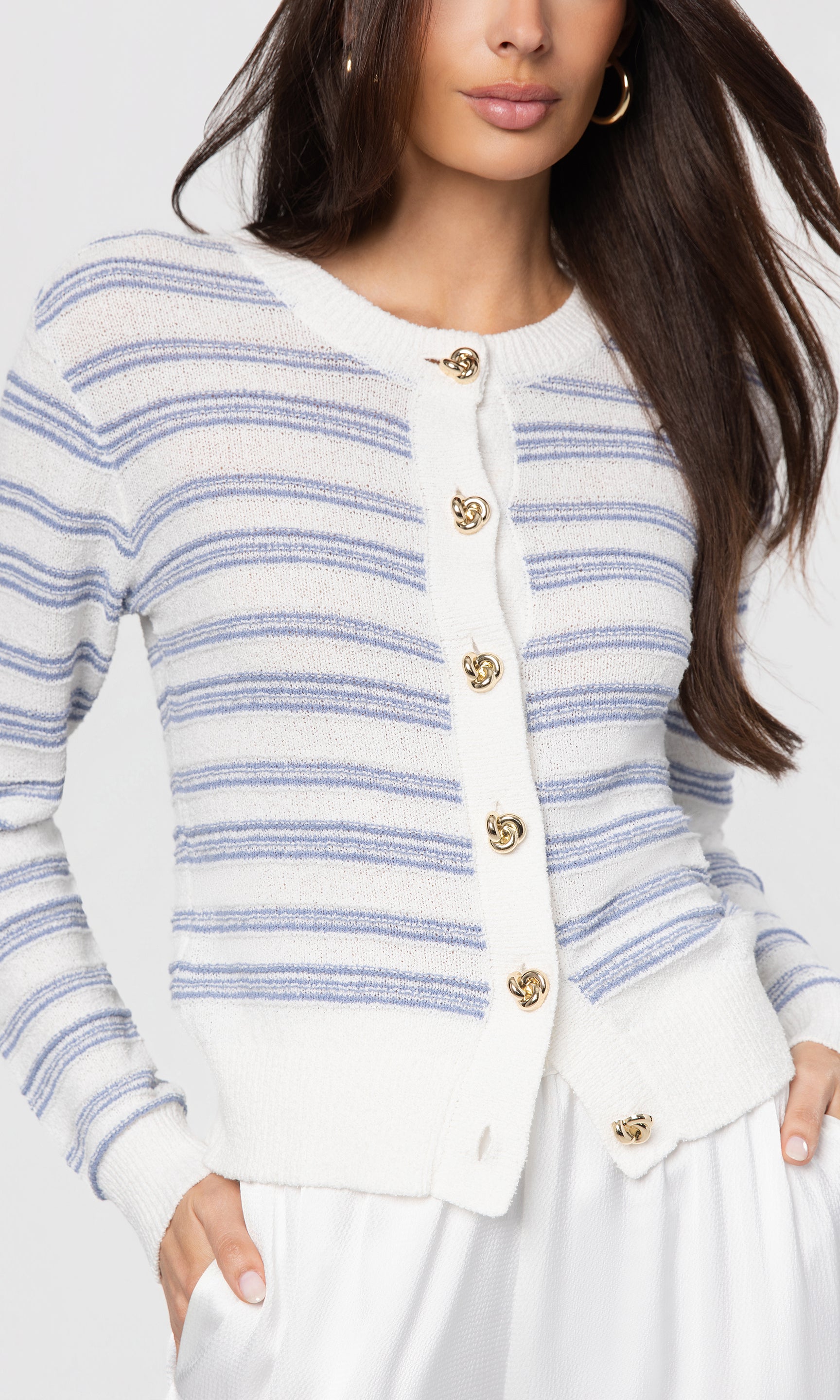 Woman wearing a white cardigan with blue stripes and gold buttons on a plain background