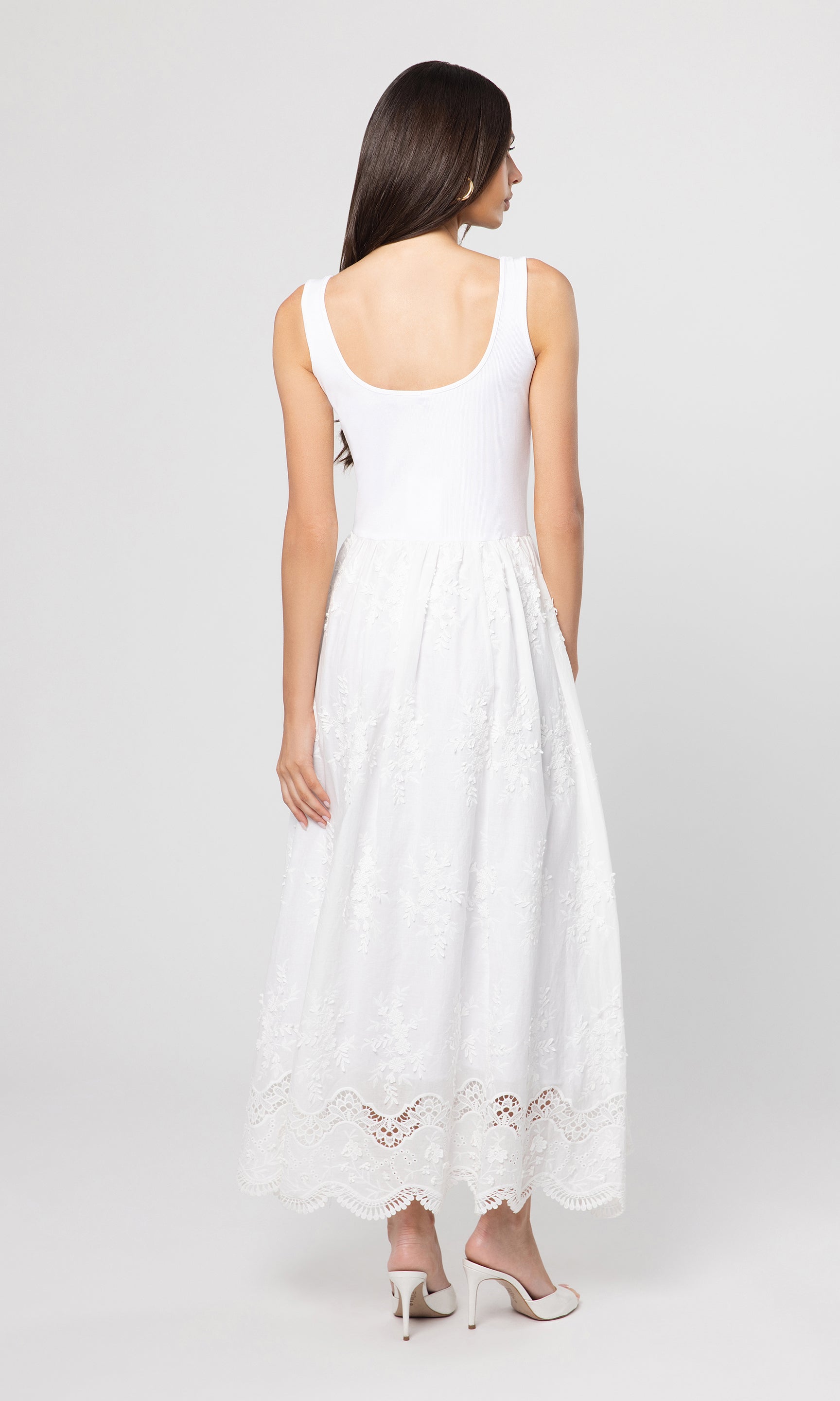 Back view of woman wearing a white sleeveless dress with lace details on a plain background