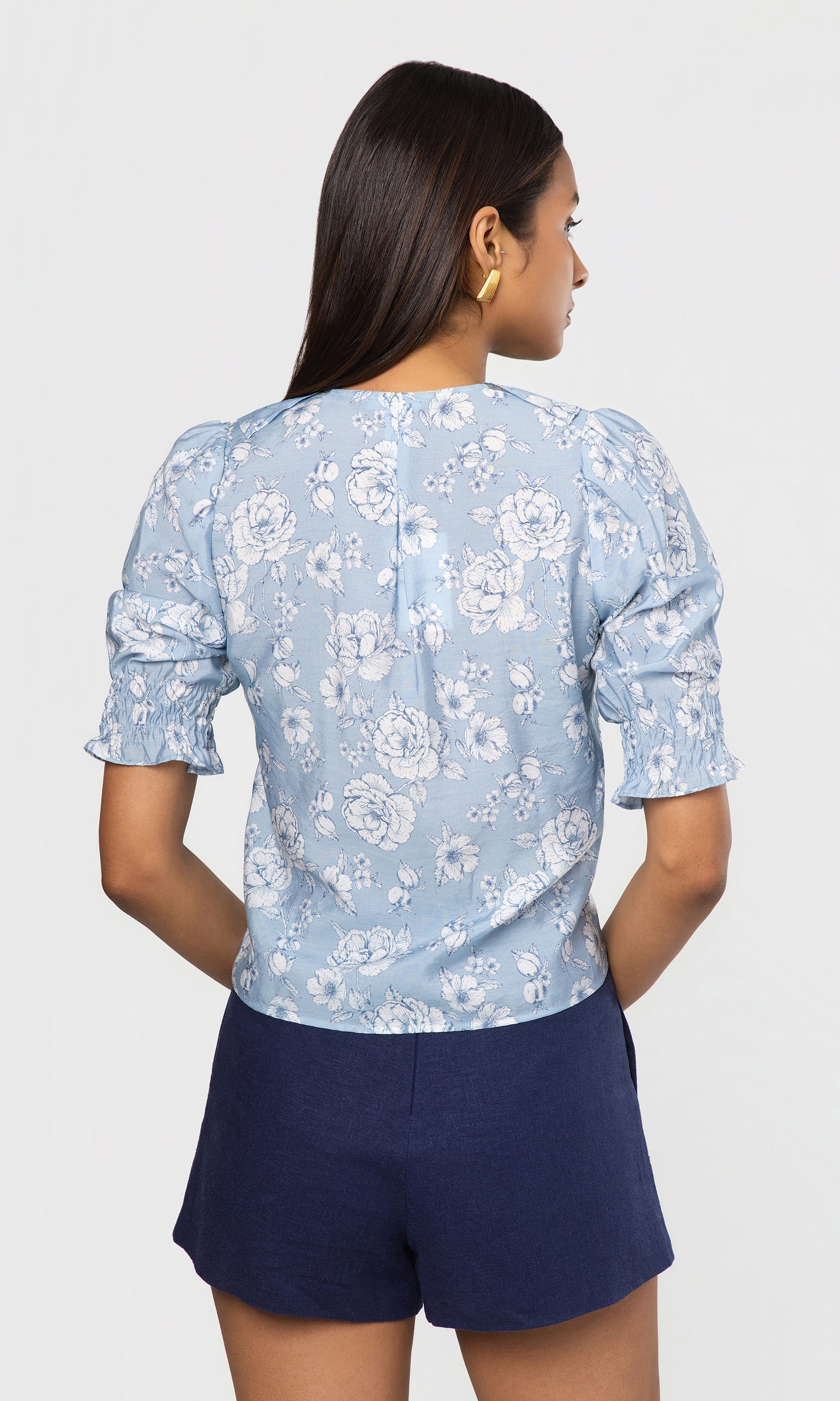 Back view of woman wearing a light blue floral blouse and navy shorts on a white background
