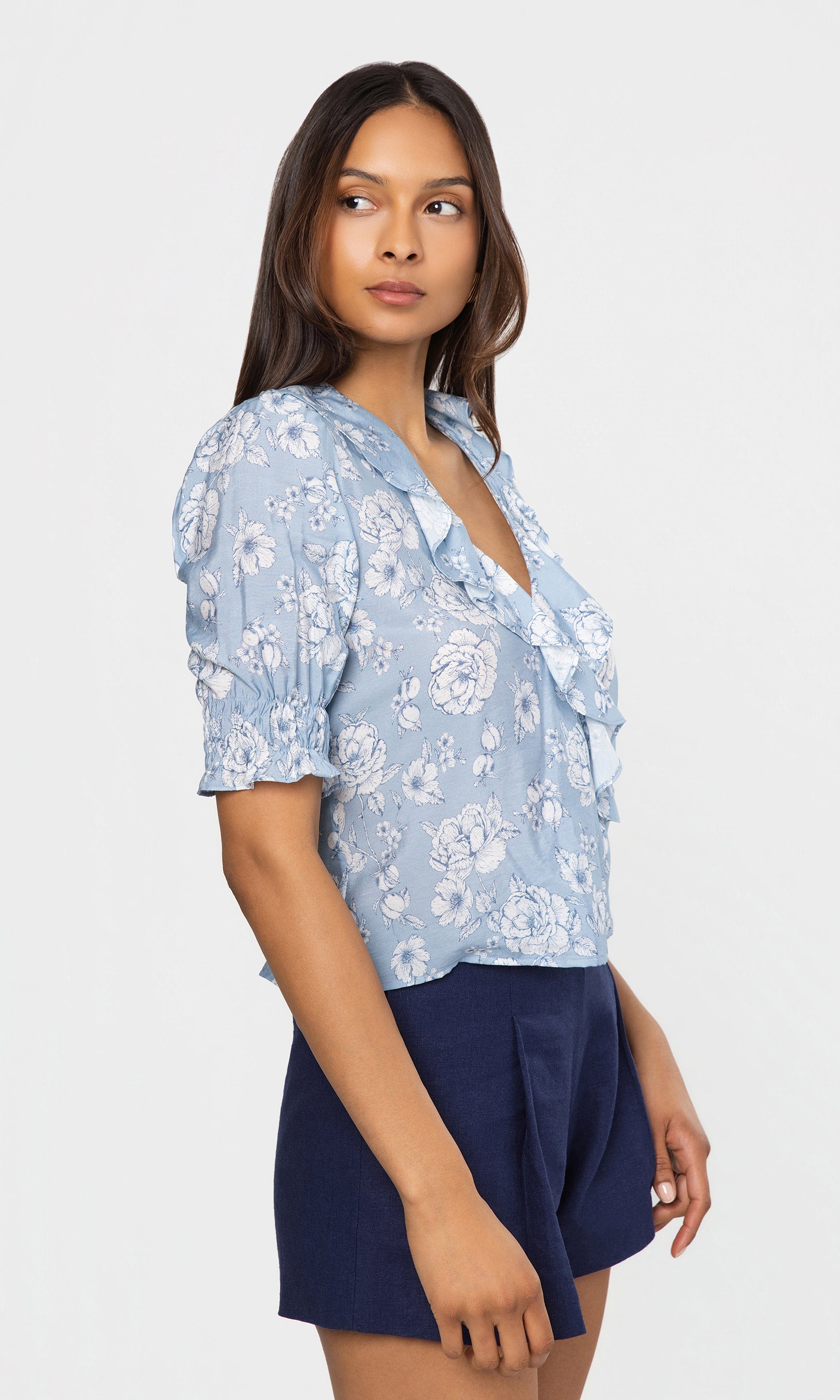 Side view of woman wearing a light blue floral blouse and navy shorts on a white background