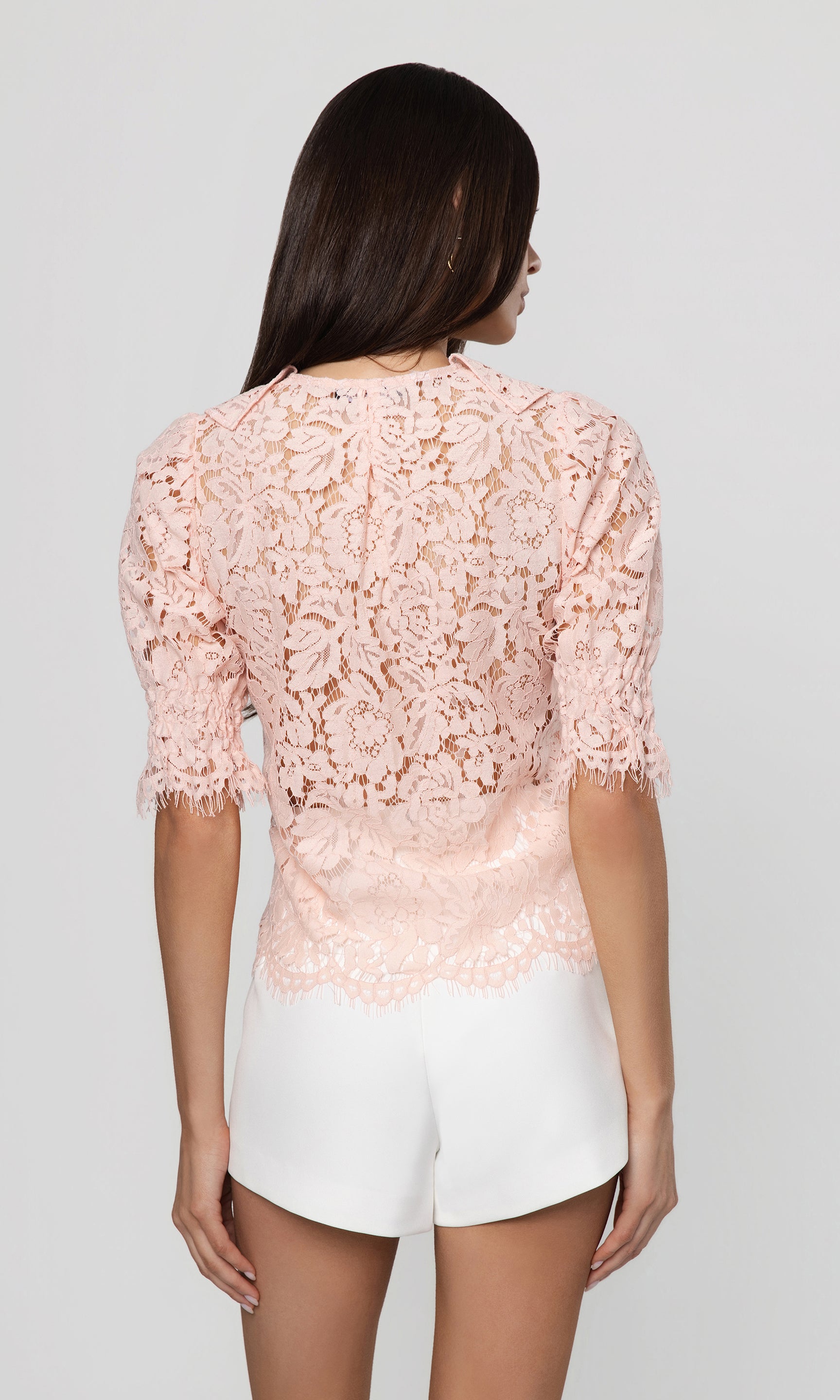 Back view of woman wearing a pink lace top and white shorts on a gray background