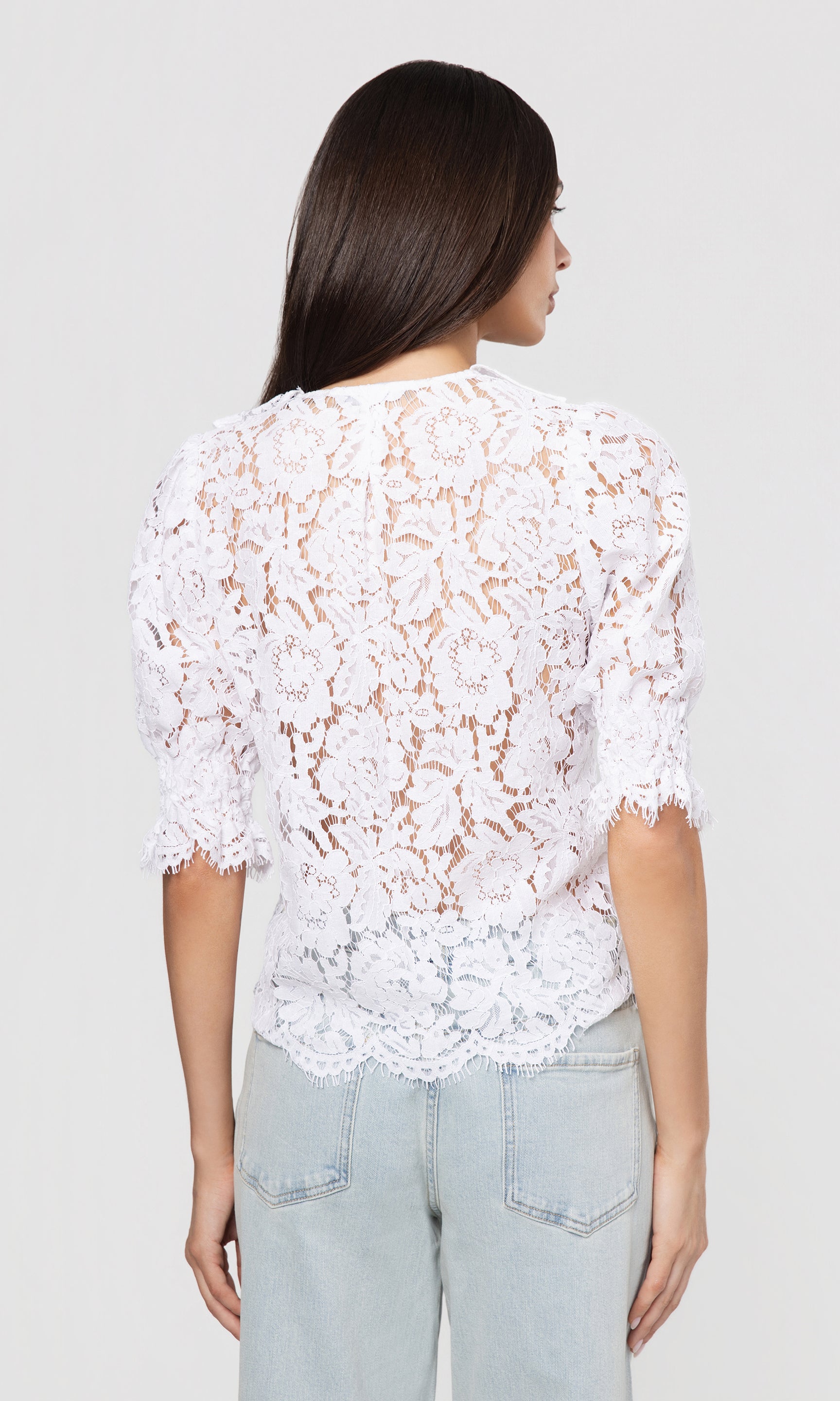 Back view of woman wearing a white lace top and light blue jeans on a plain background