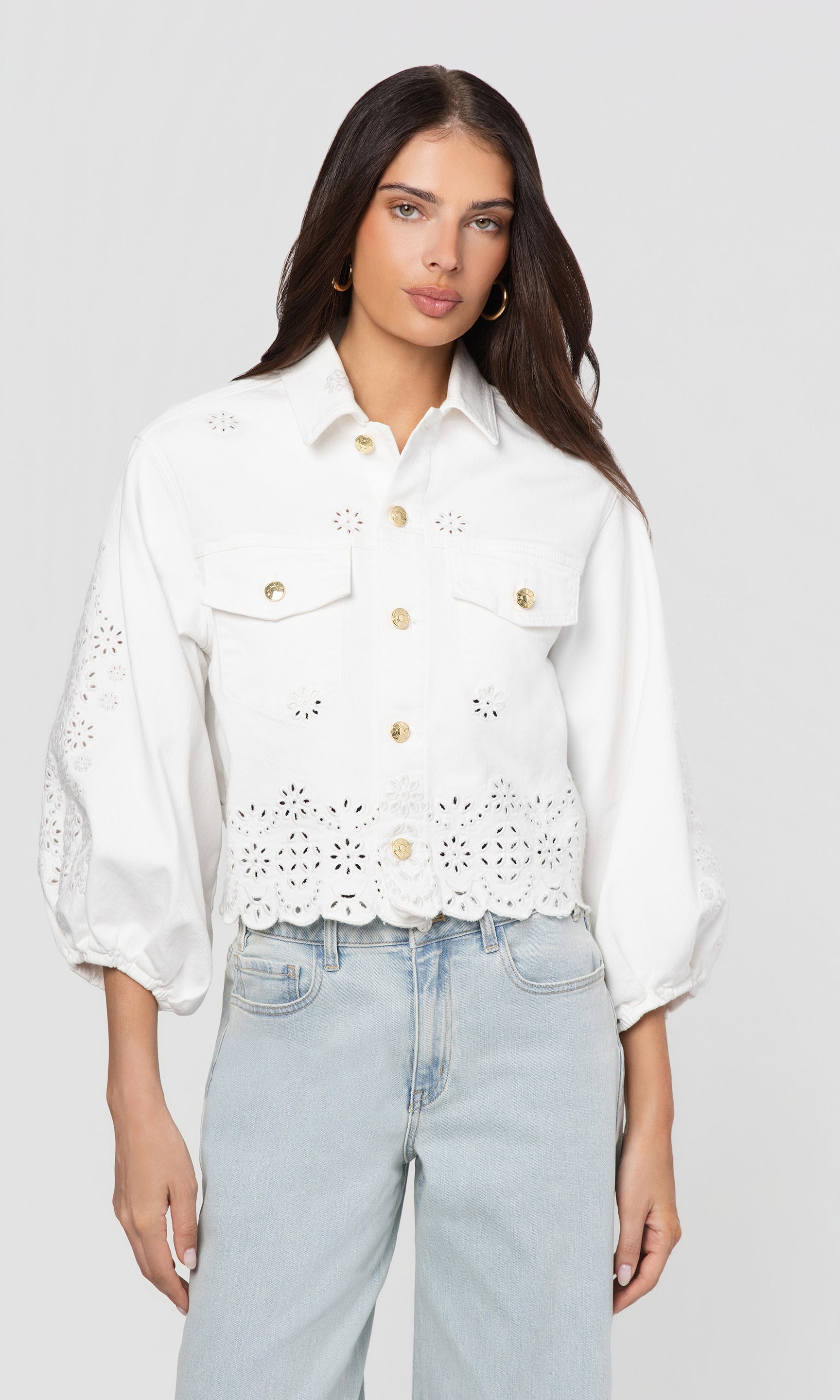 Woman wearing a white denim jacket with eyelet details and light blue jeans on a plain background