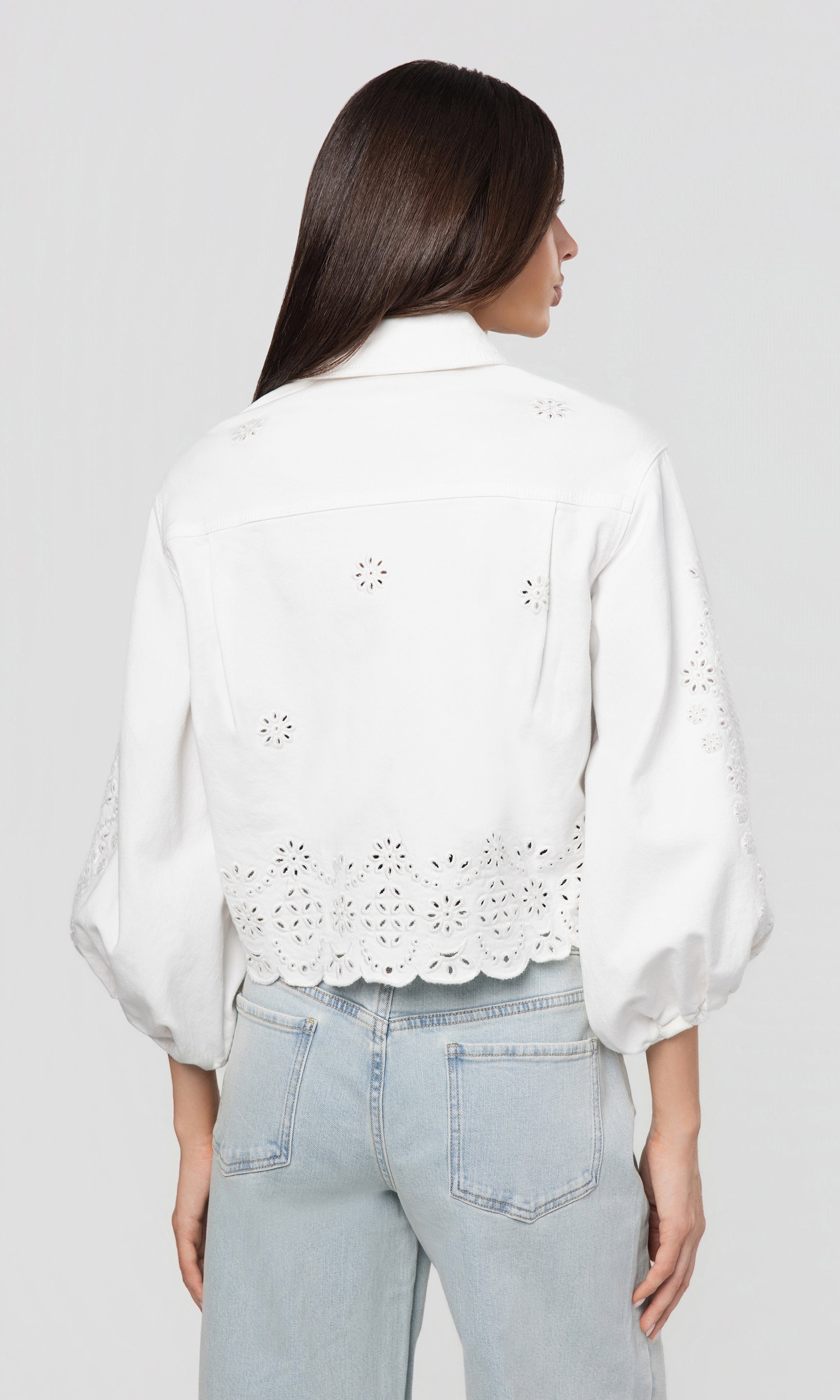 Back view of woman wearing a white jacket with eyelet details and light blue jeans on a plain background