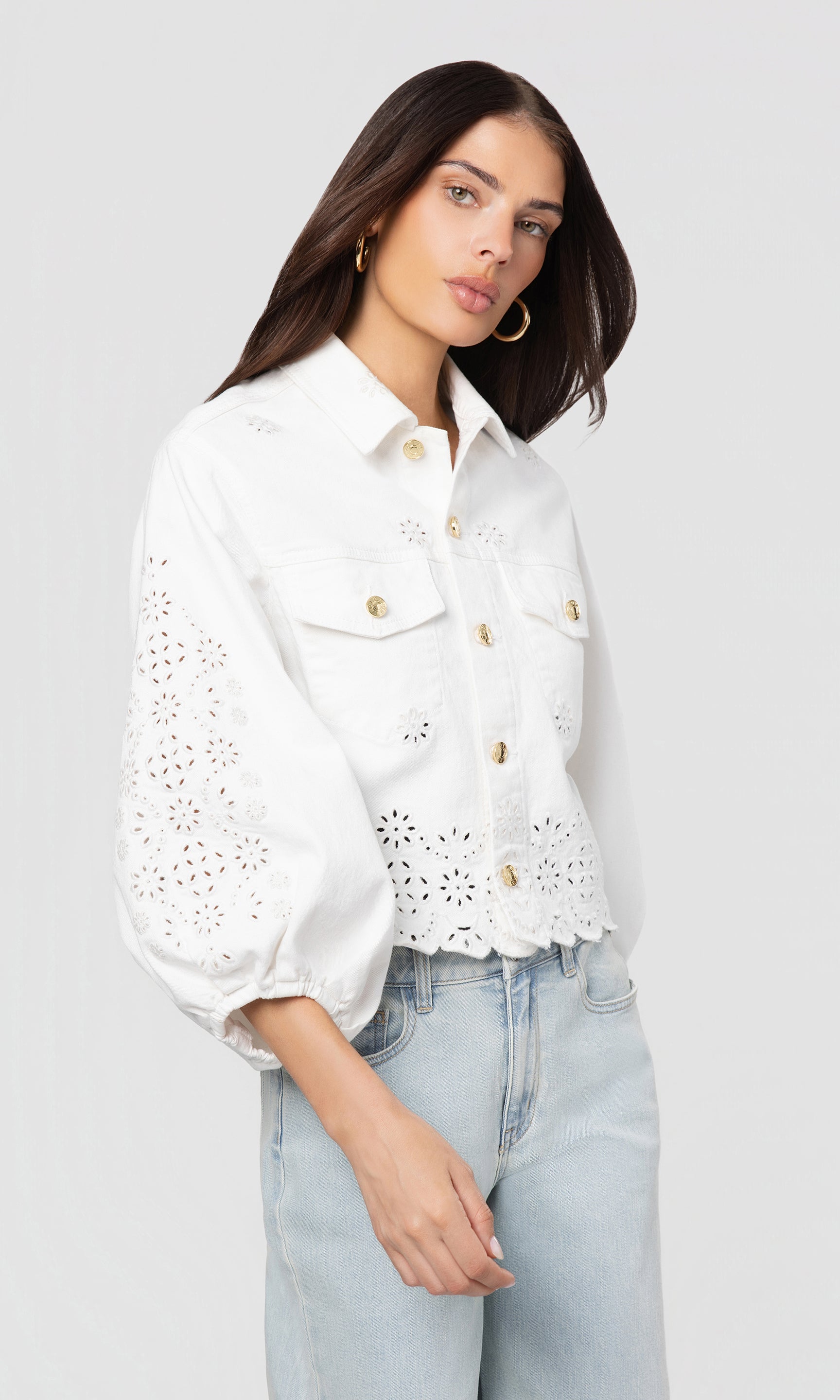 Side view of woman wearing a white eyelet blouse and light blue jeans against a plain background