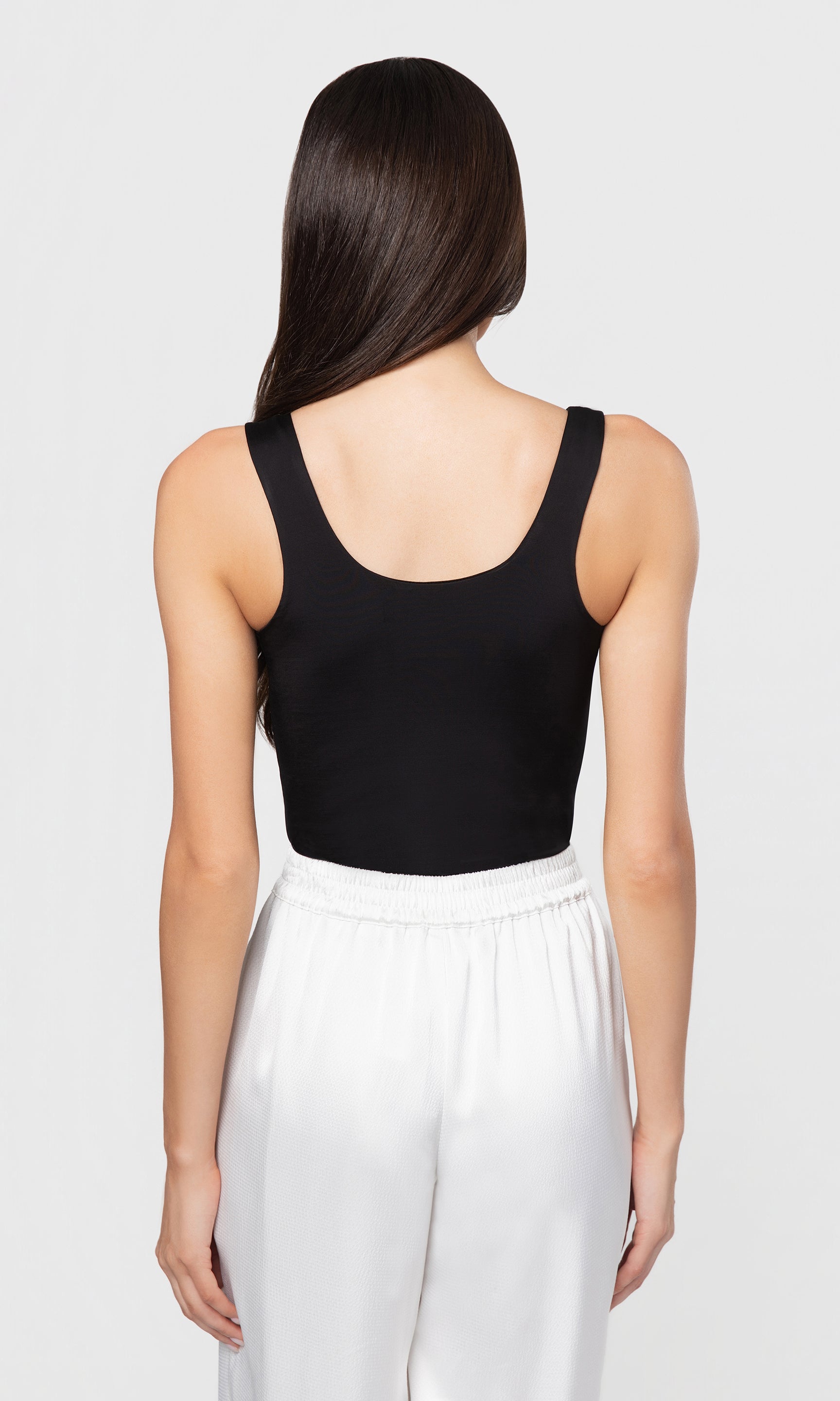 back view of person wearing a black tank top and white pants on a light gray background