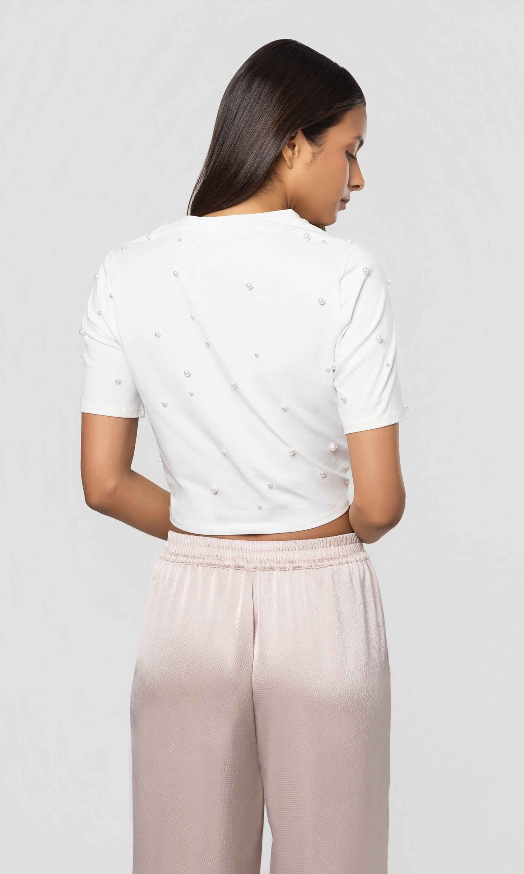 Back view of woman wearing a white top with decorative pearls and beige pants on a light gray background
